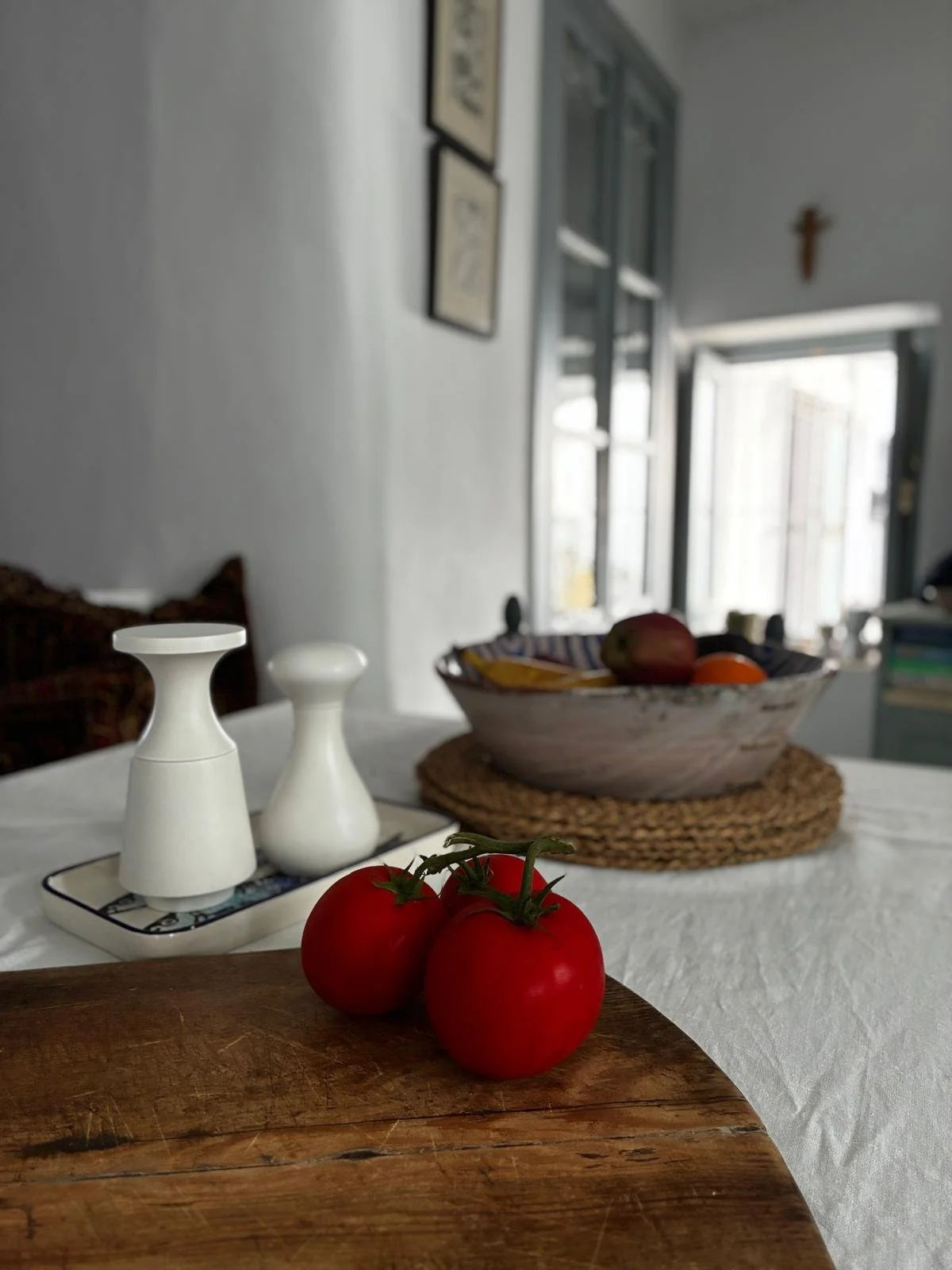 Three red tomatoes on a wooden cutting board in the foreground. In the background, a table with two white vases, a decorative tray, a bowl of fruit, and a bowl with various objects. The room has a window with open shutters and framed artwork on the wall.
