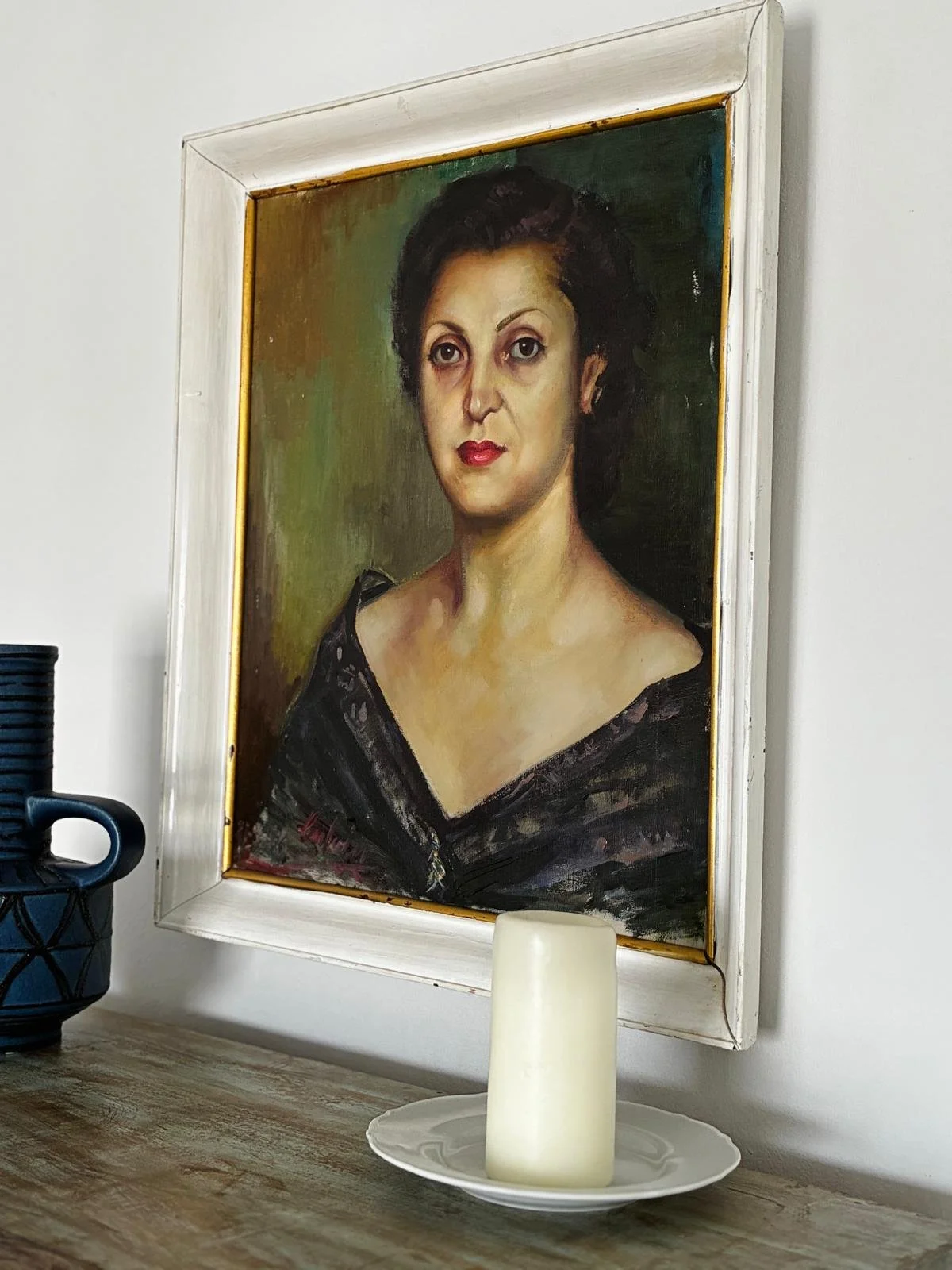 A portrait painting of a woman with short dark hair, wearing a black dress with a wide neckline, hanging on a white wall. In front of the painting on a wooden surface, there is a white candle on a white dish and a black container with a handle.