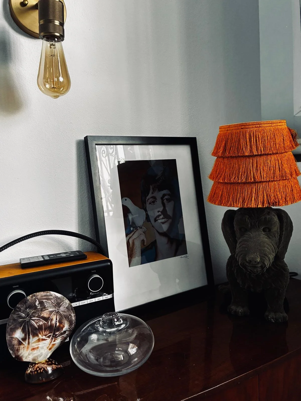 A wall decorated with a framed portrait of a man holding a bird, a vintage lamp shaped like an elephant with a ruffled orange lampshade, a framed photo, a stereo, and decorative glass objects including a glass bowl and a decorative glass palm tree figurine.