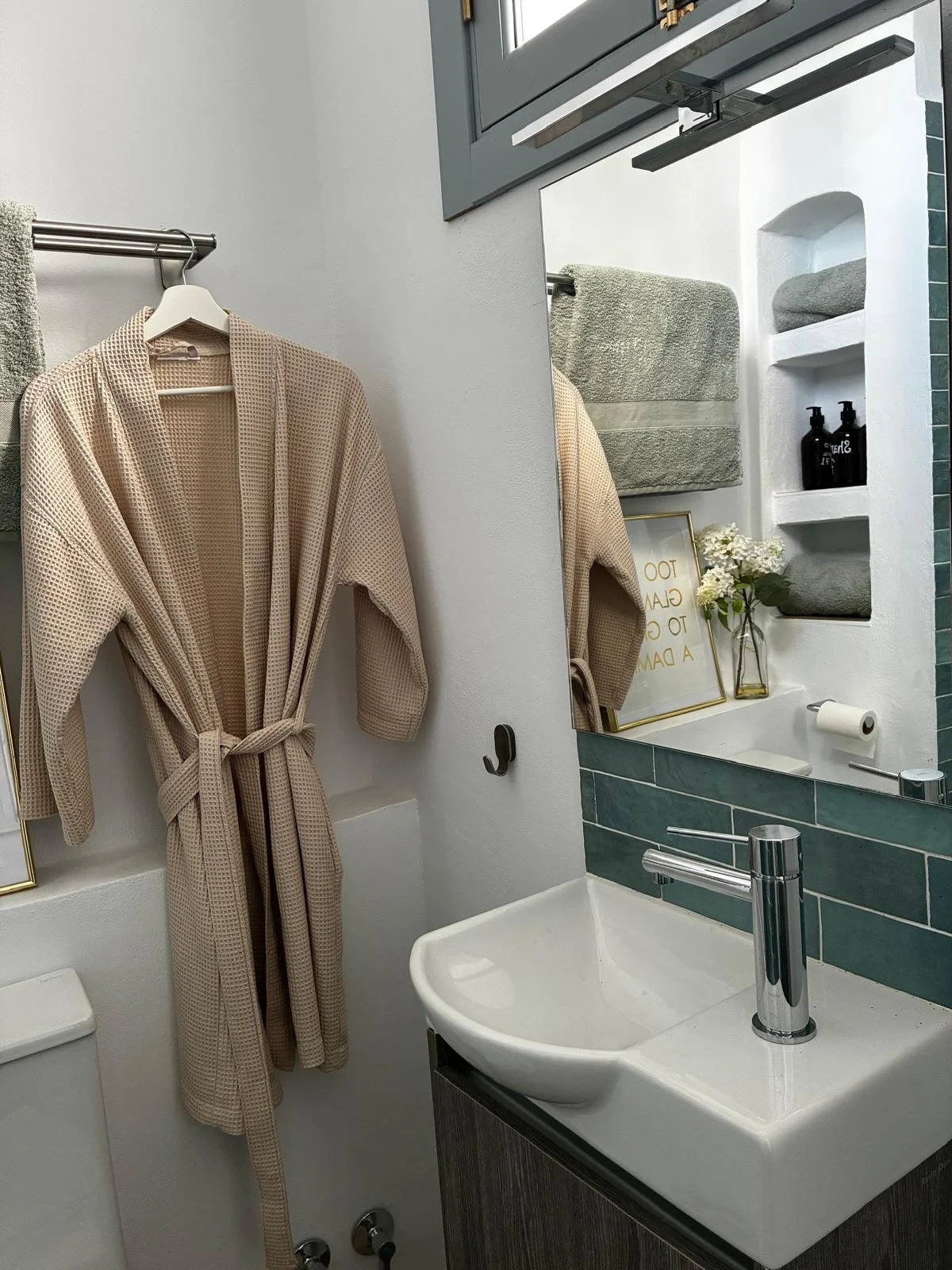 A small bathroom with a white sink, a mirror, and a gray vanity. A beige bathrobe hangs on a rack, and a towel is on a built-in shelf. A framed print and a vase with white flowers are on the shelf. There are black pump bottles and toiletries on the shelf, and a toilet paper roll is attached to the wall.