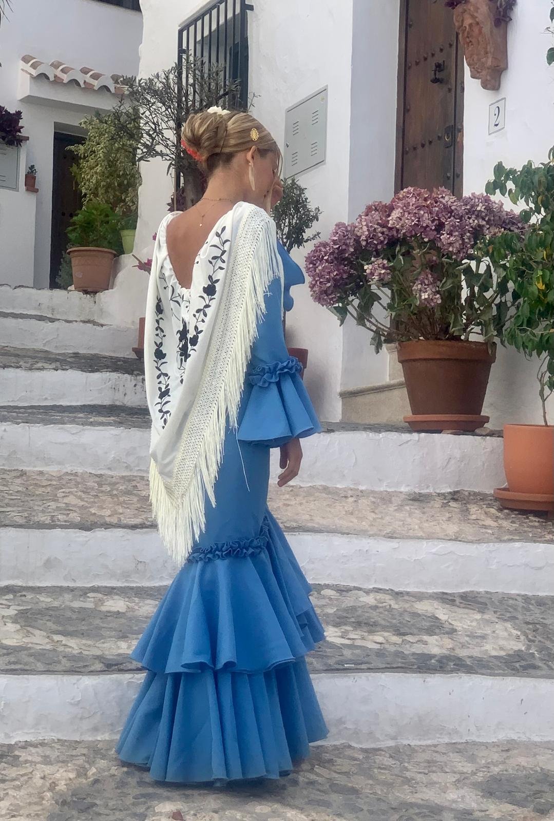 Woman dressed in a blue flamenco-style dress walking up white stairs outside a white building with potted plants.