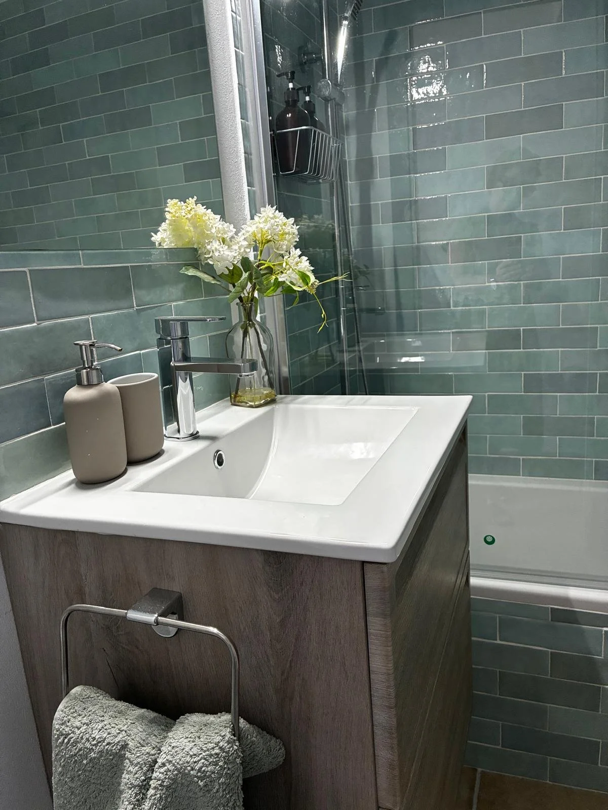 Bathroom with green subway tile wall, white sink, wooden cabinet, beige soap and lotion dispensers, glass vase with white flowers, and a towel rack with two rolled towels. Shower with glass door and shelves, holding dark soap bottles.