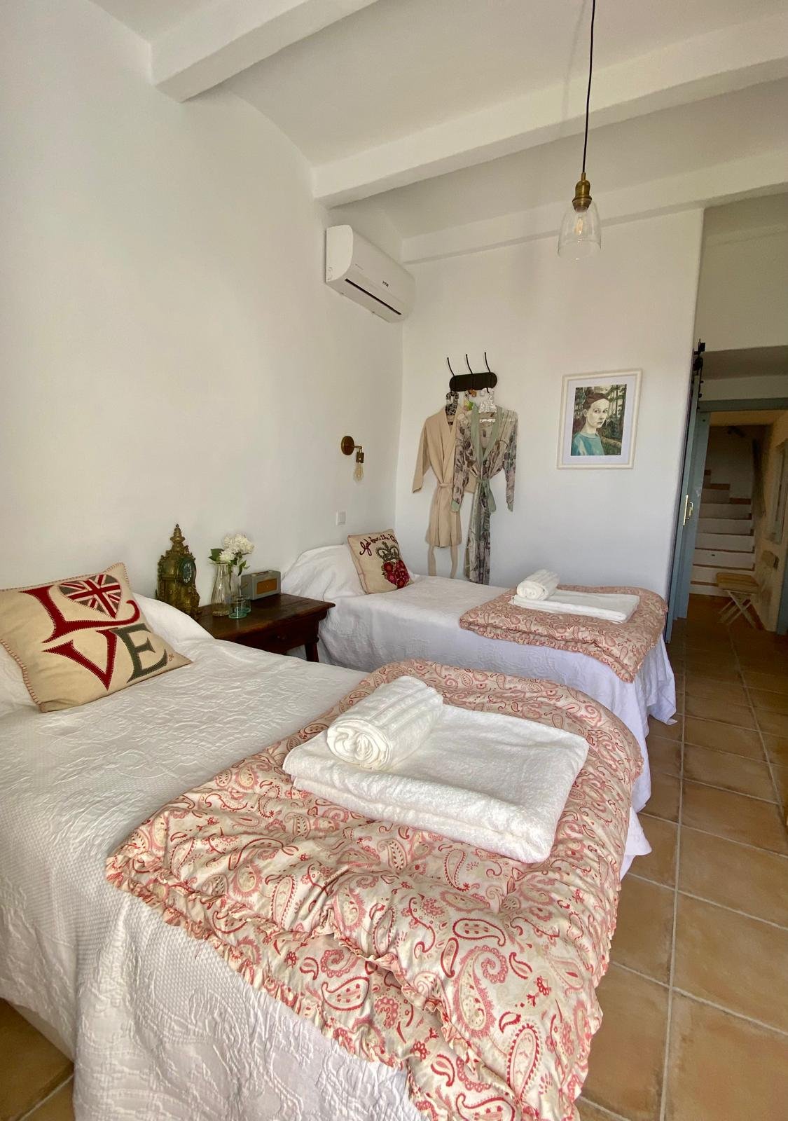 A bedroom with two single beds, each covered with white bedspreads and pink patterned throws, fold-out towels, a pillow with a love design, and decorative pillows. There is a small bedside table with a clock, flowers, and an ornament. The wall has hanging clothes and a framed portrait. The room has terracotta tile floors and a hanging light fixture.