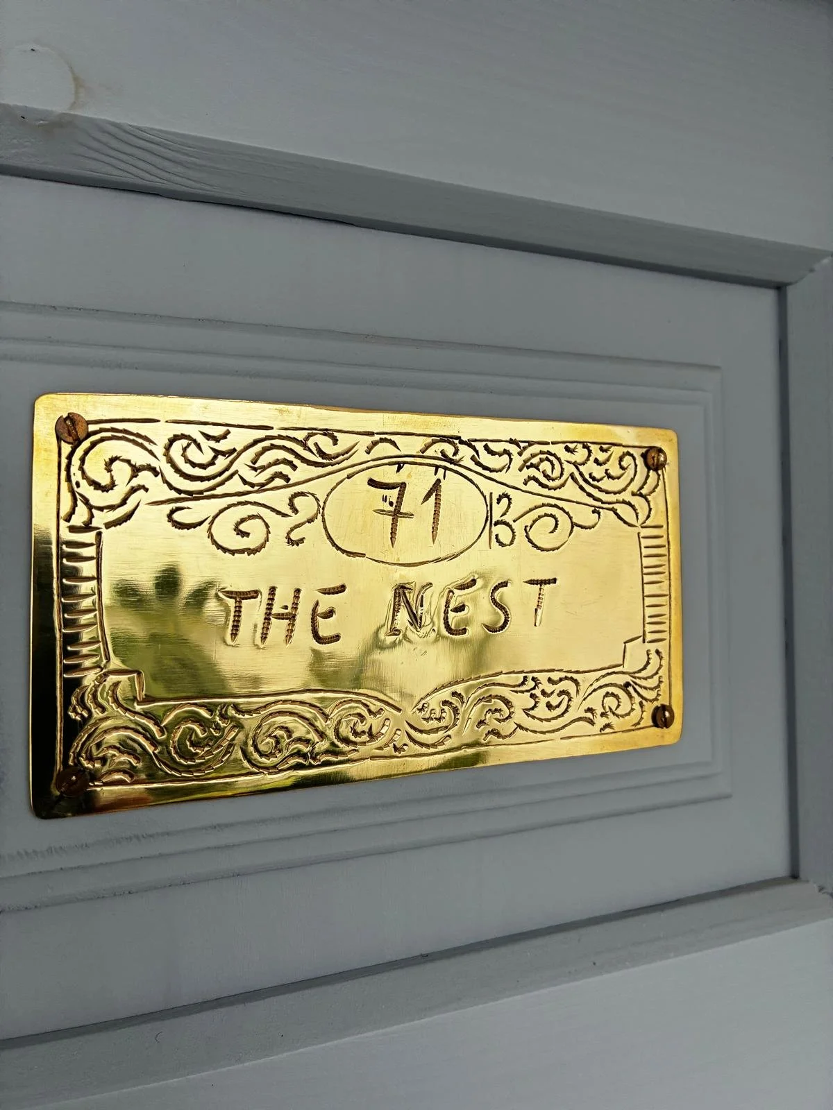 A gold-colored plaque with an ornate design, with the words Número 71 and THE NEST engraved on it, mounted on a light grey wall.
