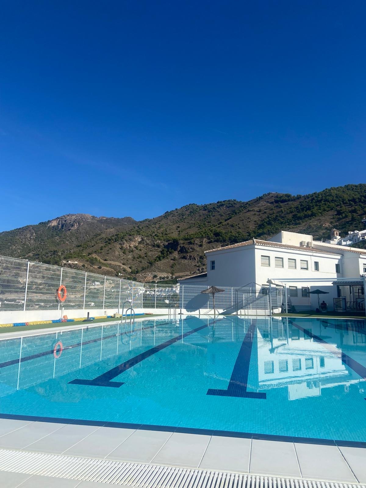 Outdoor swimming pool in Frigiliana with mountain view and white building that contains a cafe in the background, under a clear blue sky.