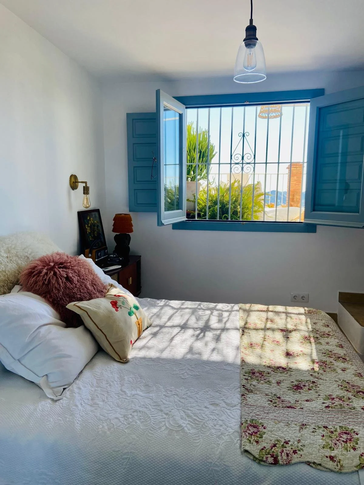 A bedroom with a bed, floral quilt, pillows, a nightstand with a lamp and a framed picture, and an open window with blue shutters. Outside, there are plants and trees visible through the window, with natural daylight illuminating the room.