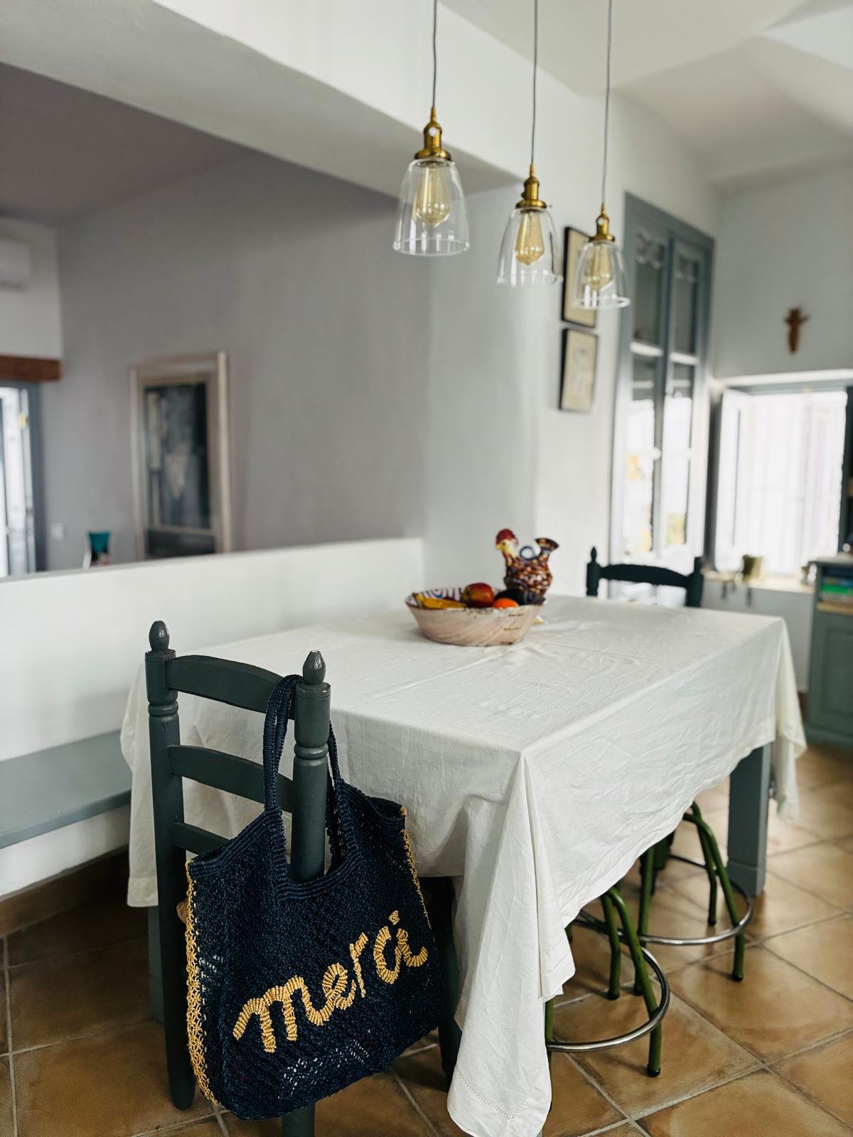 A dining room with a white tablecloth-covered table, black wooden chairs, a basket with fruits and a colorful rooster figurine, and a black tote bag hanging on one of the chairs. There are hanging pendant lights above, an open window, and decorative items on the walls.