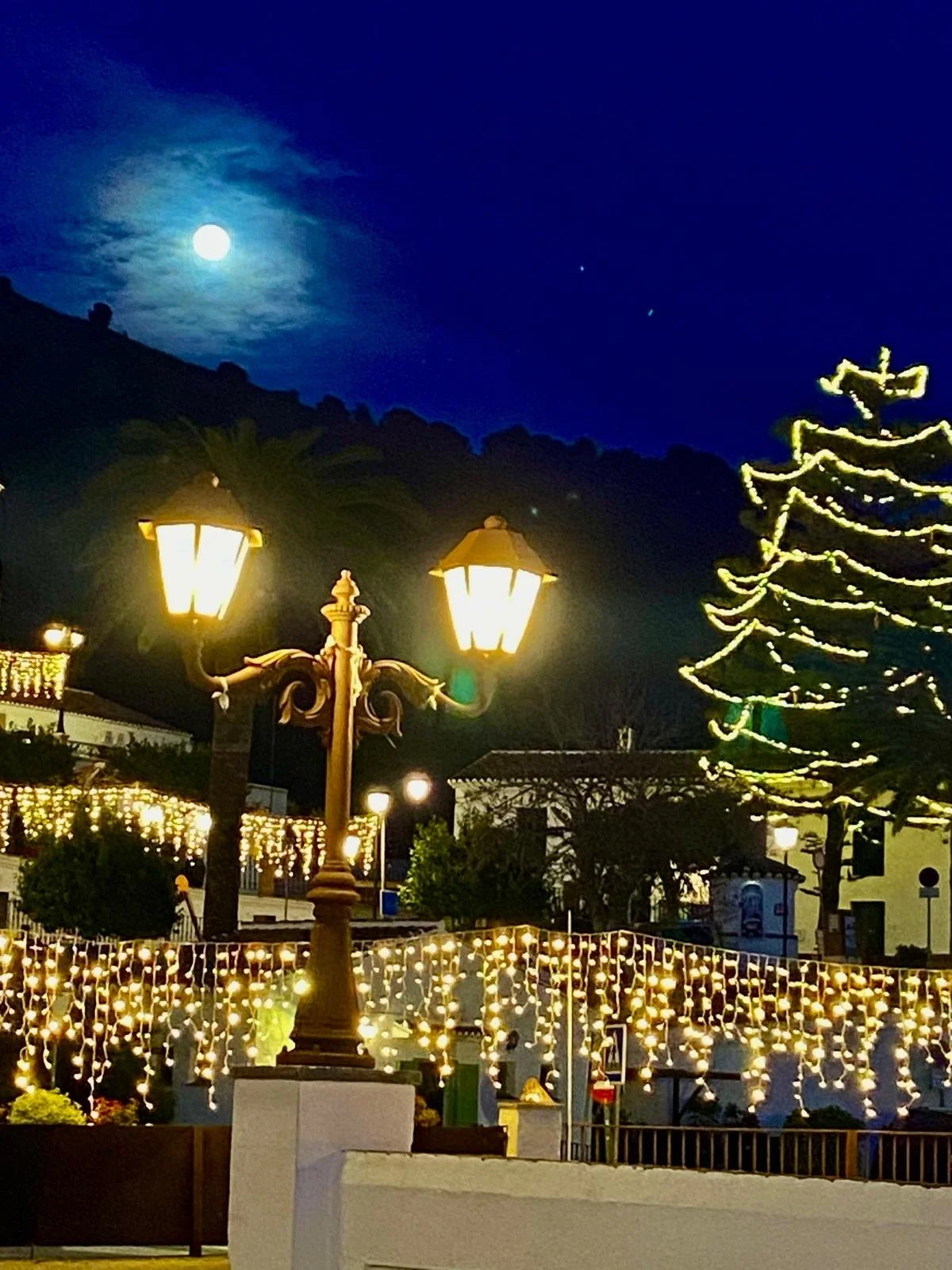 Nighttime scene with a bright full moon in a partly cloudy sky, illuminated street lamps, a decorated Christmas tree with lights, and a festive outdoor setting with string lights and people.