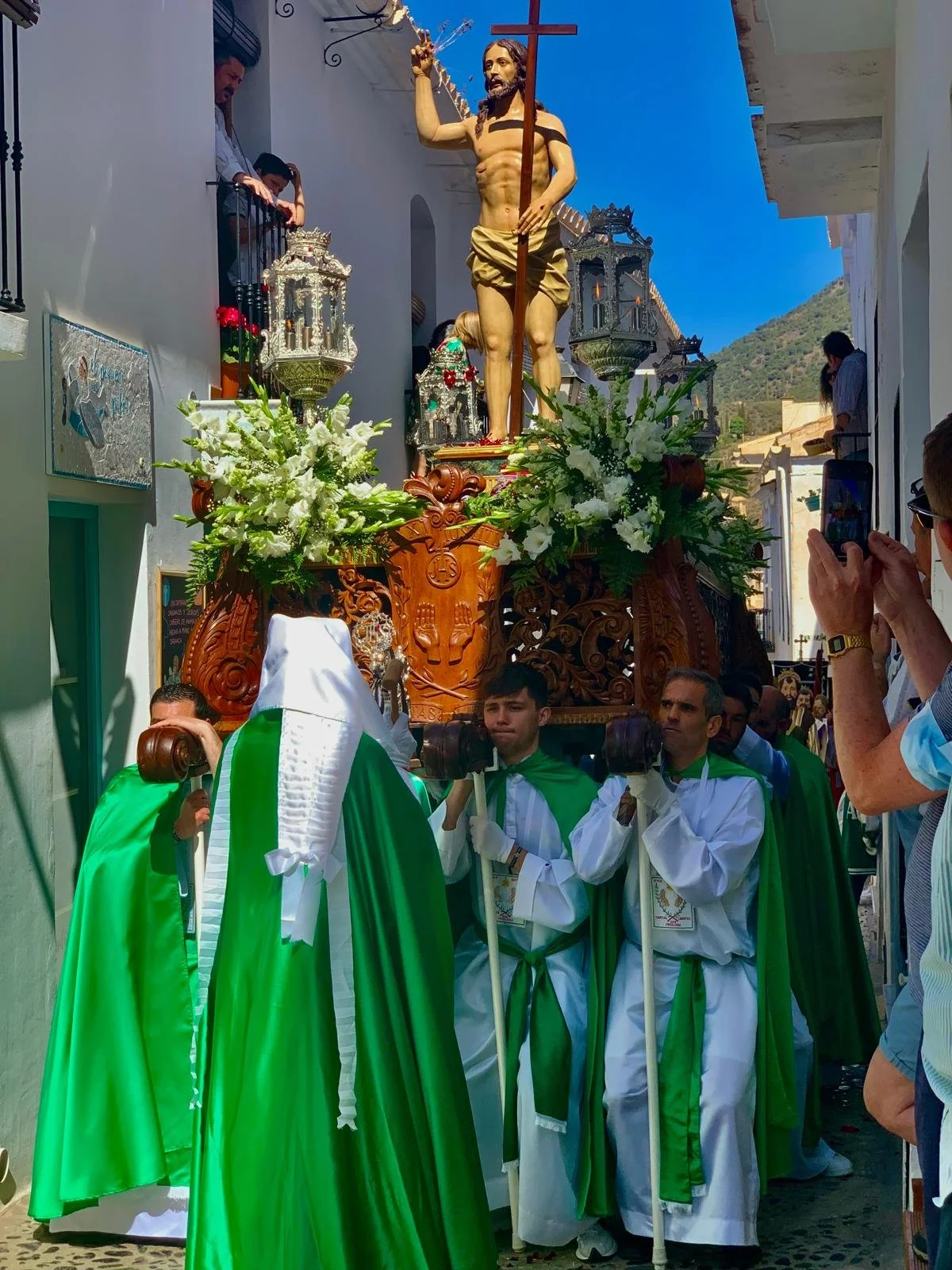 Men dressed in white robes with green cloaks carry a religious float featuring a statue of Jesus Christ holding a cross, decorated with flowers and lanterns, during a procession.