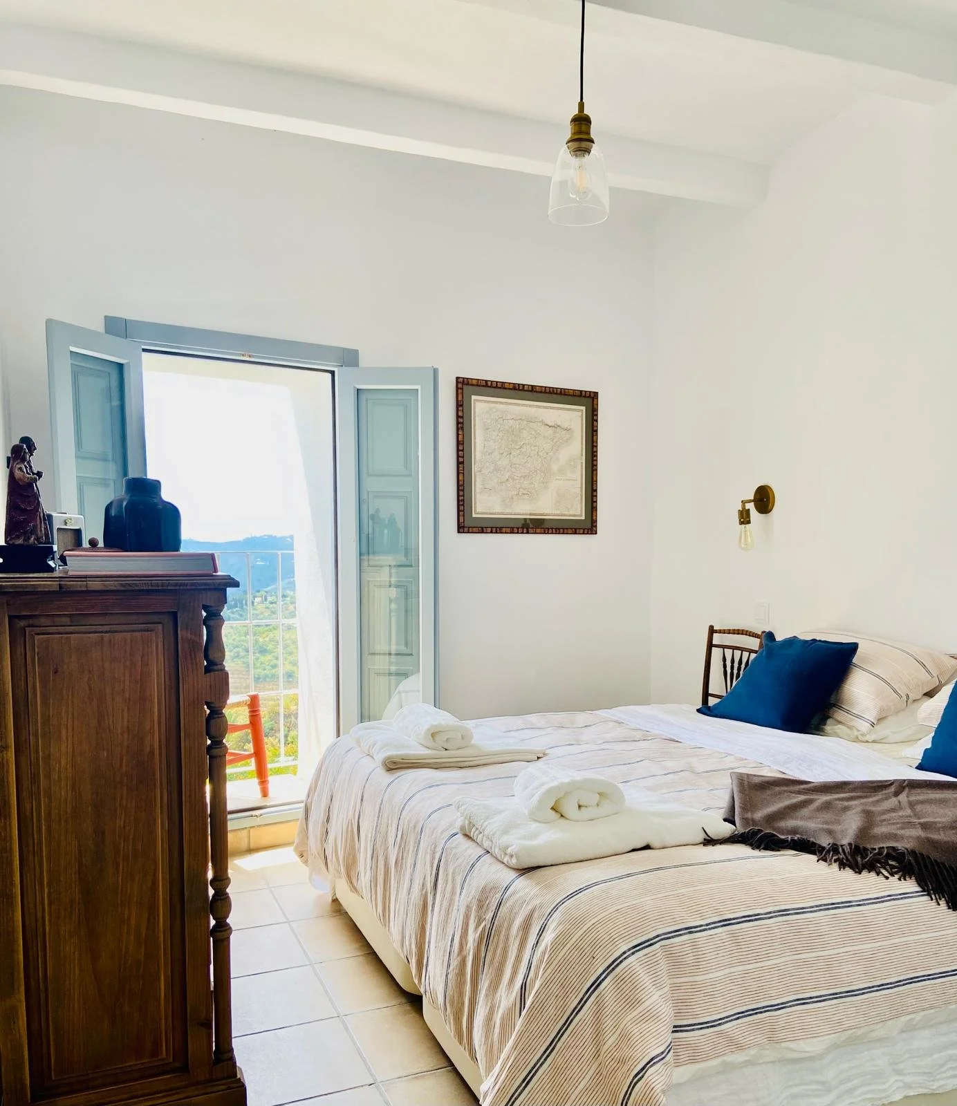 A bright bedroom with a large bed made with striped linen, blue pillows, and white towels. An open balcony door with blue shutters reveals a scenic view. There is a wooden dresser with decorative items and a wall-mounted light fixture.