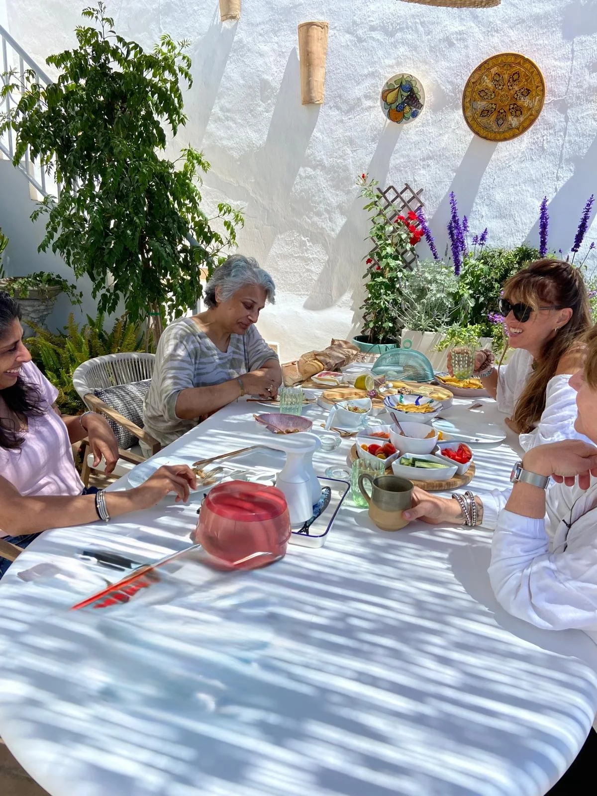 Group of women enjoying a meal at a table outdoors decorated with colorful bowls of vegetables, bread, and drinks, surrounded by lush green plants and decorative wall hangings.