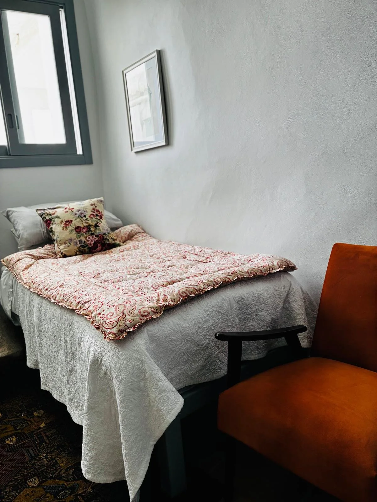 A small bedroom with a bed covered in a white quilt and pink paisley blanket, a floral pillow, a window with blue trim, a framed picture on the wall, a brown armchair with black armrests partially visible, and a patterned carpet.
