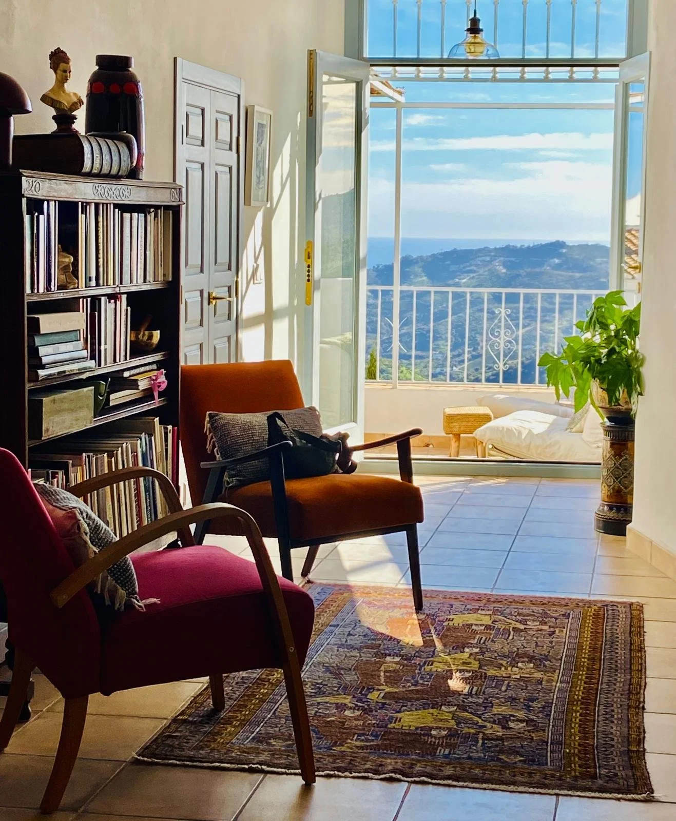 Living room with bookshelves, multiple chairs, and an open door leading to a balcony with a view of hills and the ocean.