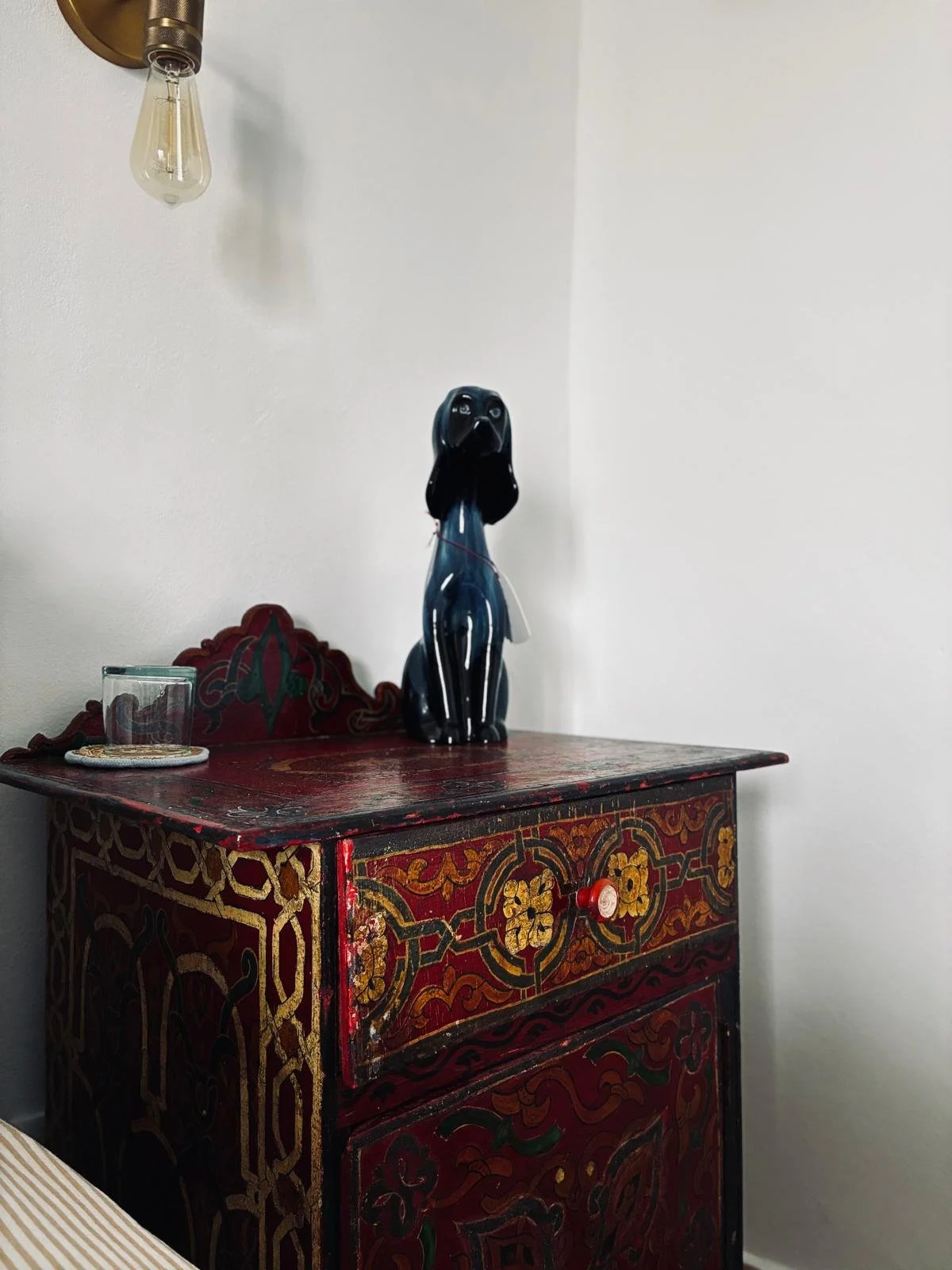 Decorative antique wooden cabinet with red, yellow, green, and black intricate floral and geometric patterns, a black dog-shaped ceramic figurine on top, a glass and coaster, and a wall-mounted vintage light fixture.