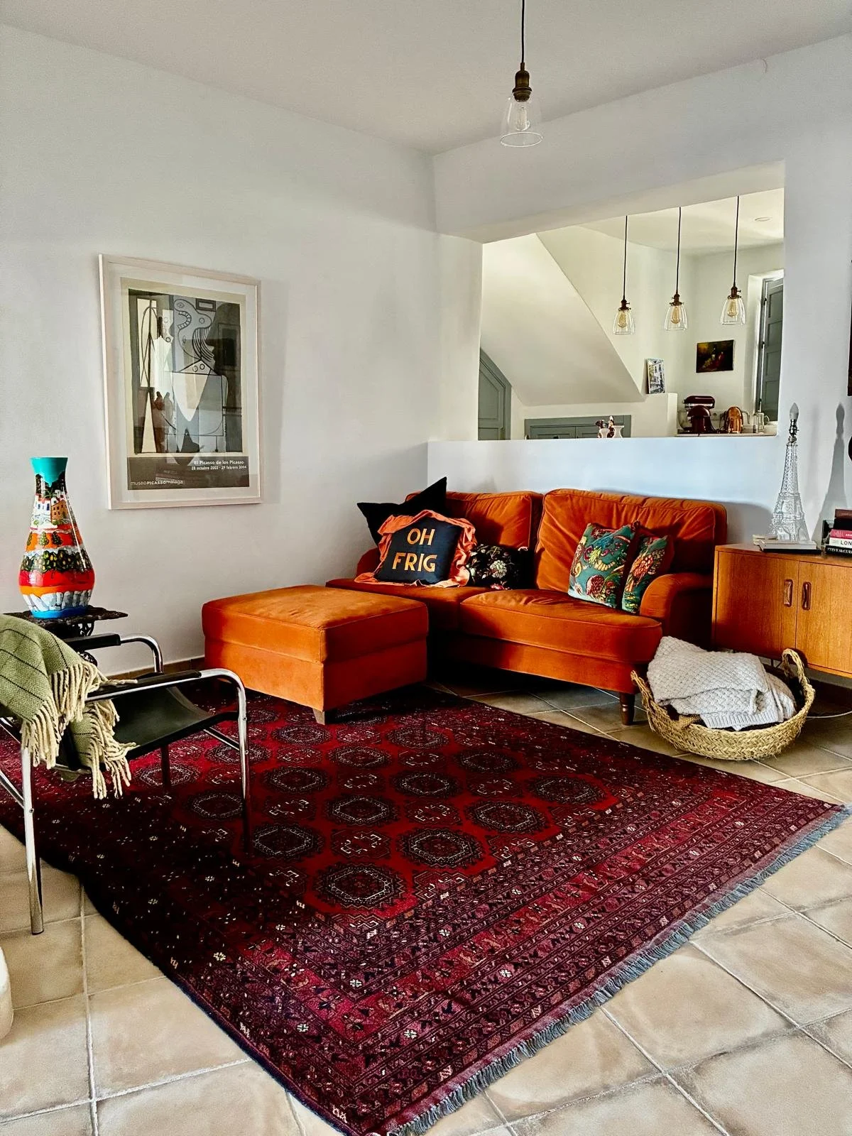 Living room with orange sectional sofa, decorative pillows, a red Persian rug, a round wicker basket with blankets, a colorful vase, a black chair with green blanket, framed wall art, a wooden cabinet, and hanging pendant lights.