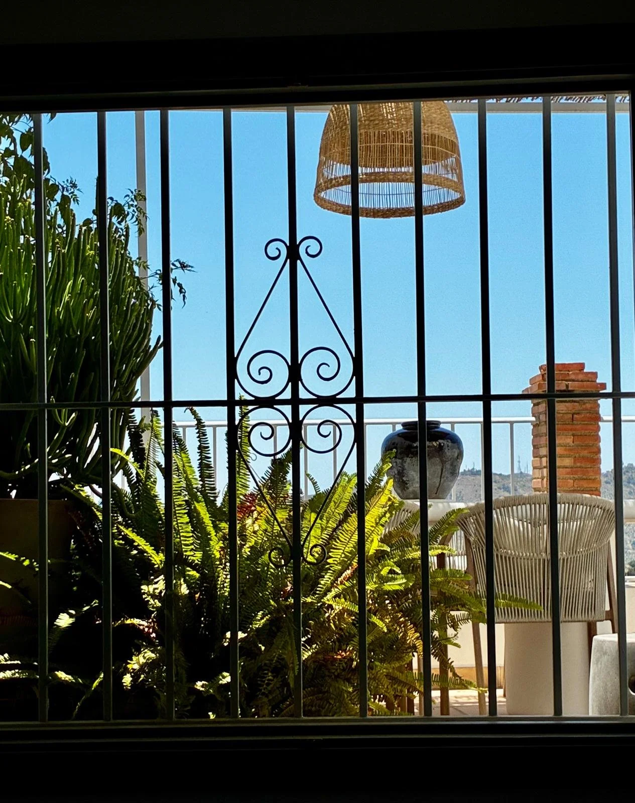 View through a window with black metal bars, showing a backyard with lush green plants, a large black and white ceramic vase, a wicker chair, and a brick chimney against a clear blue sky.
