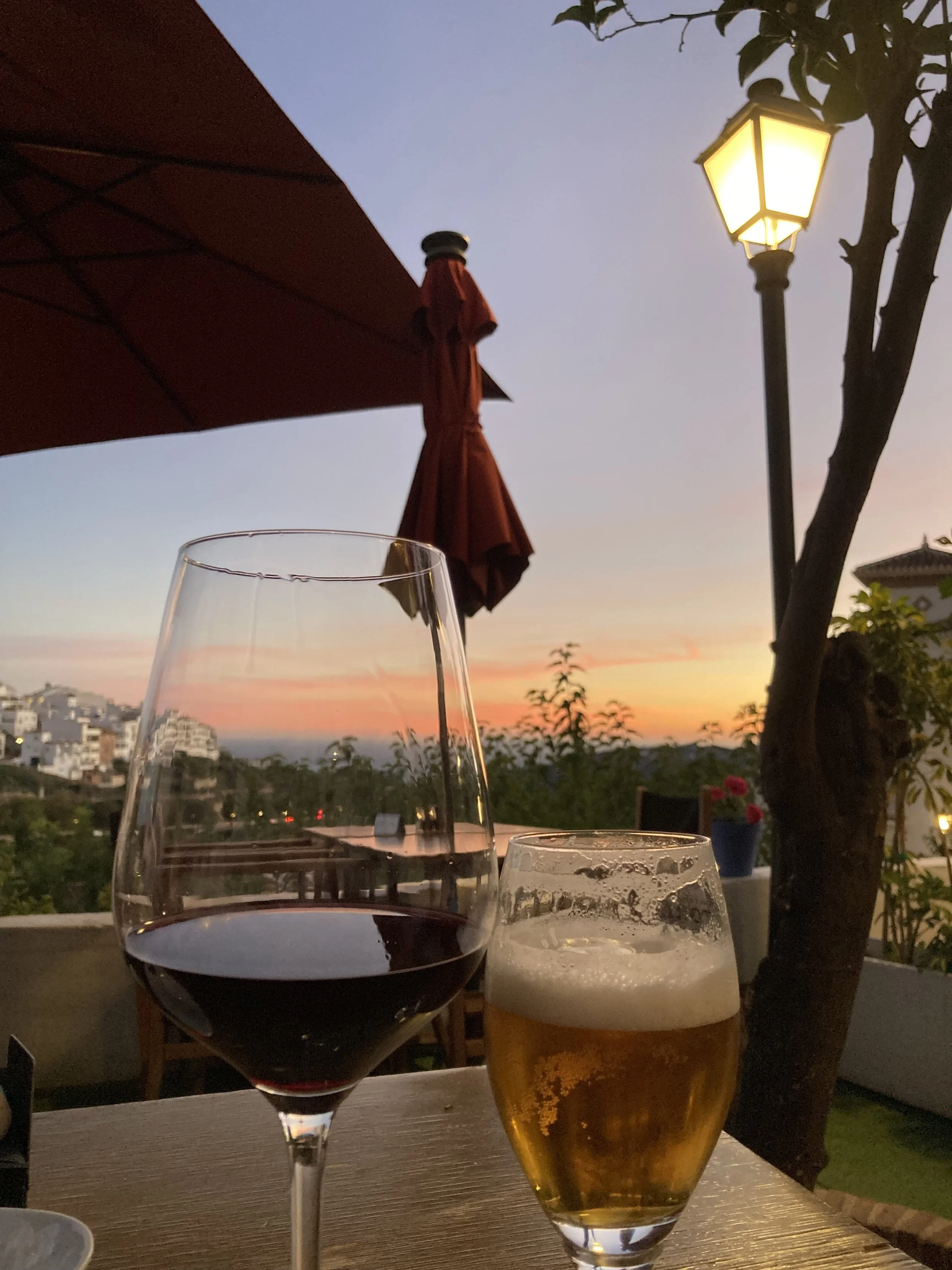 A glass of red wine and a glass of beer on a table outdoors during sunset, with a lit streetlamp, and a background of a hilly landscape and a colourful sky at sunset.
