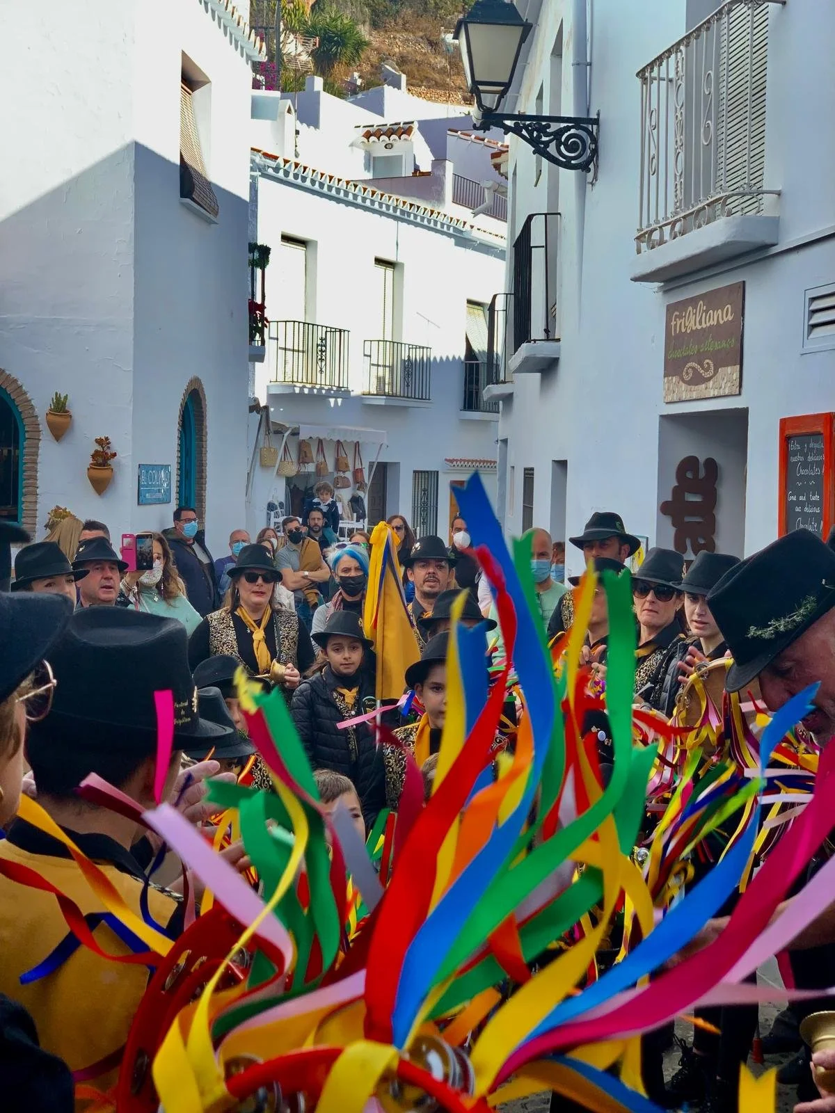 People gathered in a street during one of the festivals in Frigiliana, many wearing black hats, some with masks, surrounded by colourful ribbons and decorations, with white buildings in the background.