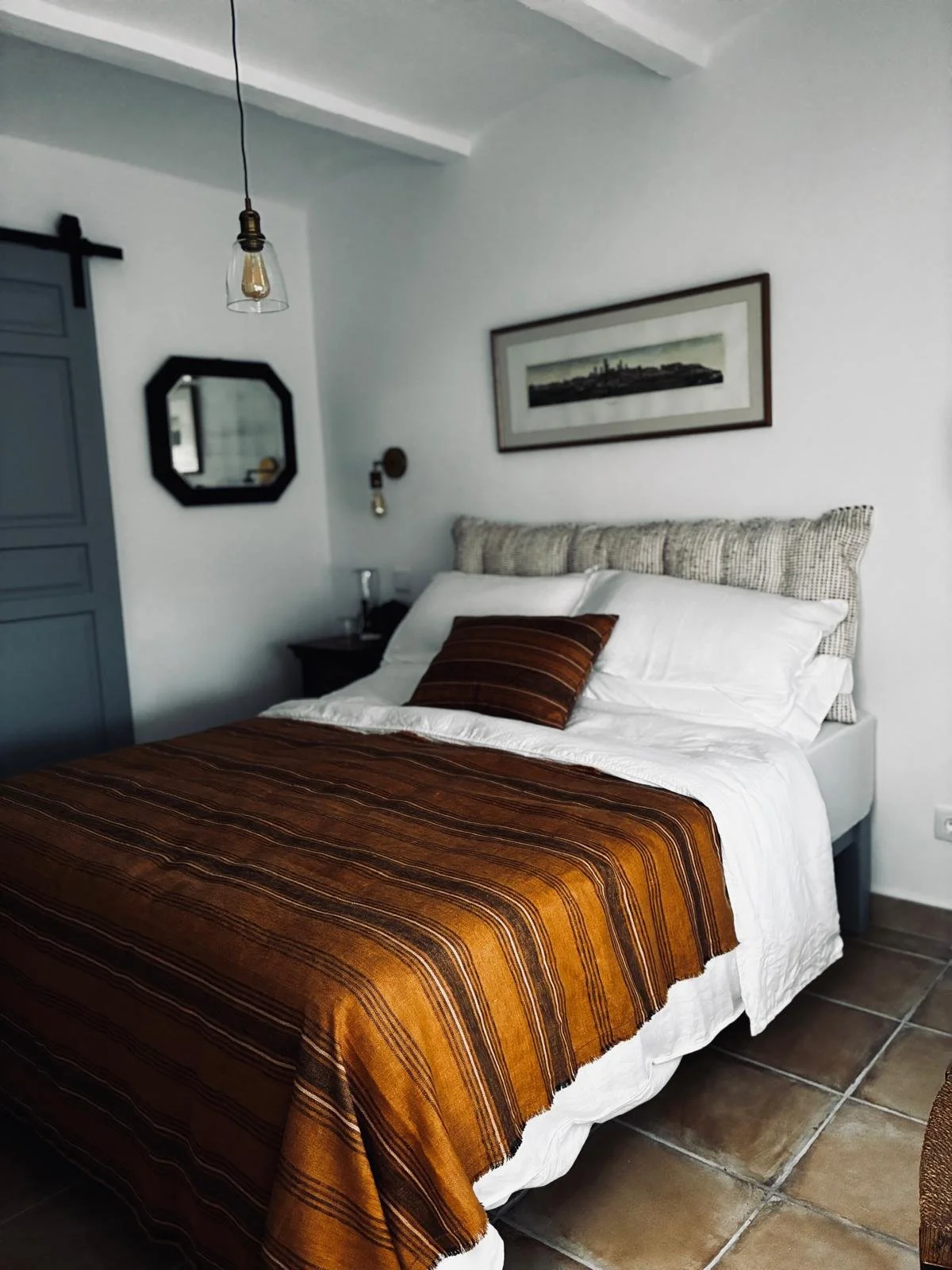A neatly made bed with white sheets, a brown striped blanket, and matching pillow, in a modern bedroom with white walls, a framed landscape picture, a small nightstand with a glass, a hanging light bulb, and decorative mirror.