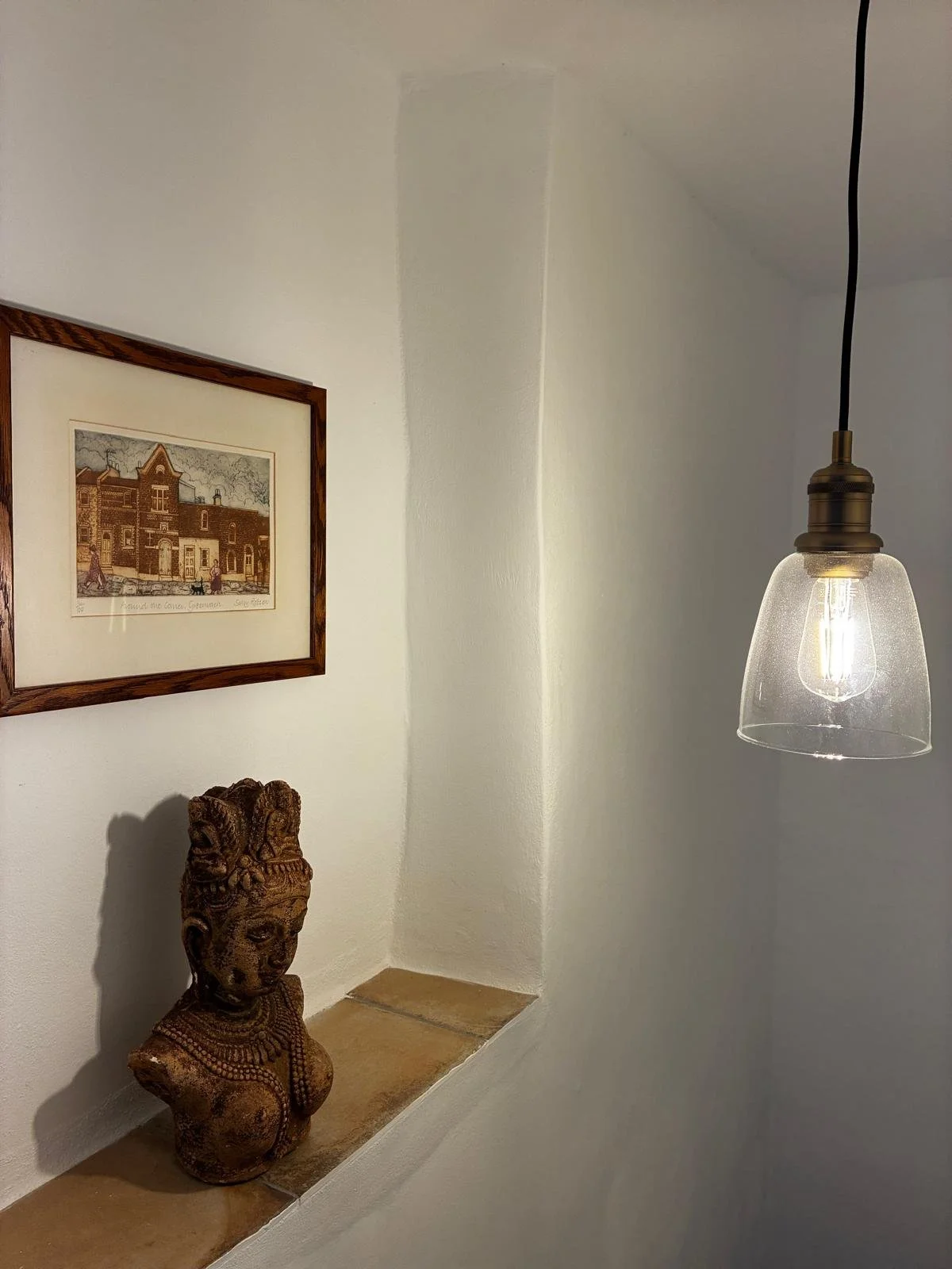 Decorative wall corner with framed picture of a building, a wooden carved bust, and a hanging light fixture with a clear glass shade and Edison bulb.