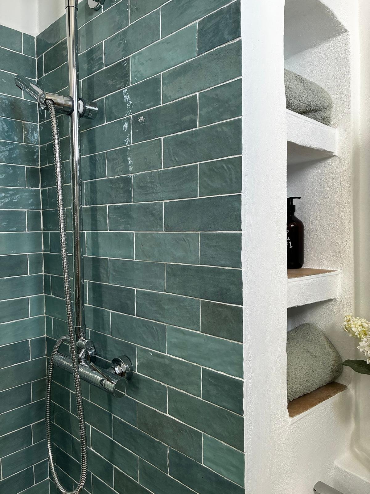 A shower area with green rectangular tiles on one wall and a white wall with built-in shelves holding a soap dispenser, rolled towels, and decorative items.
