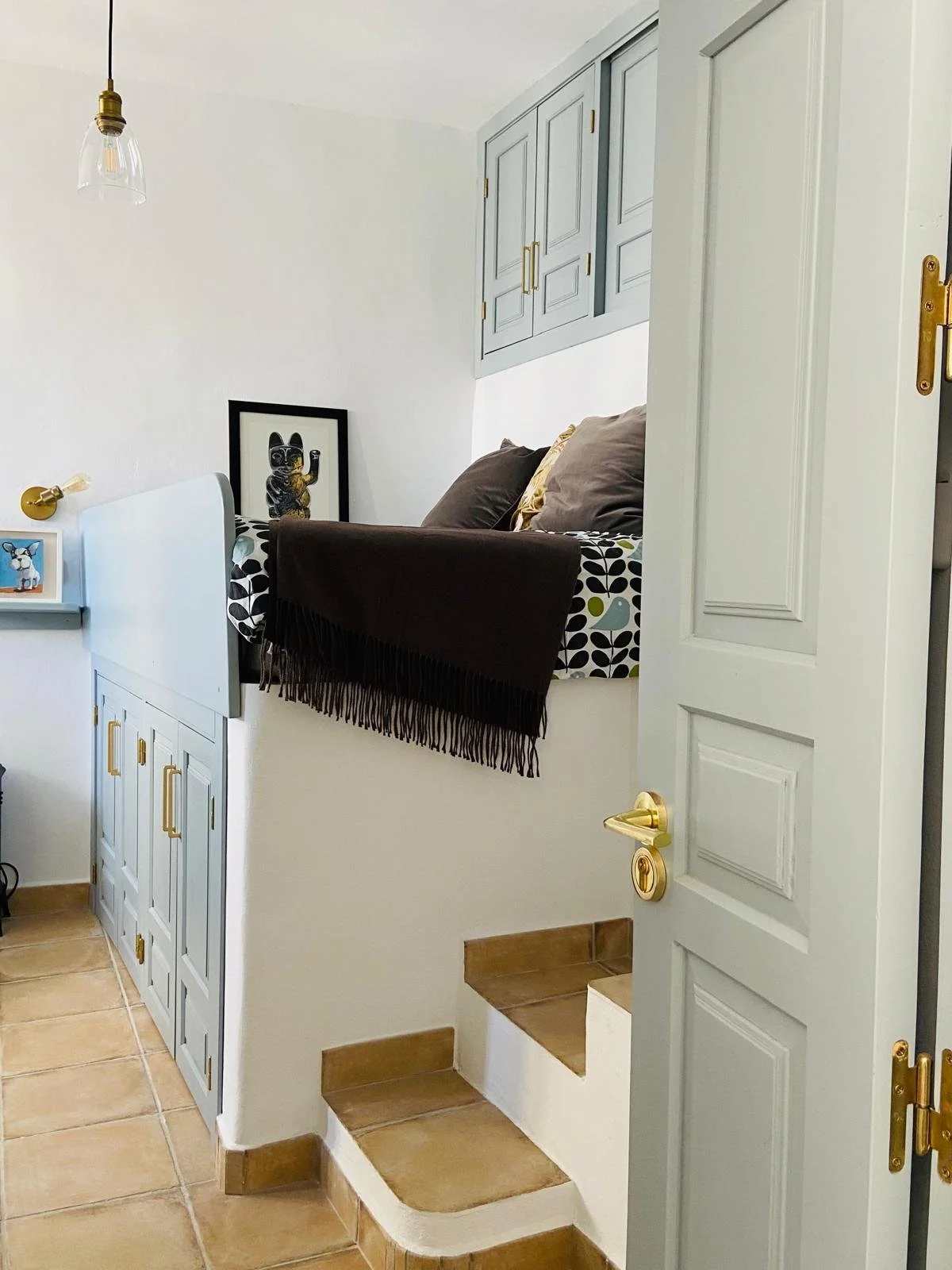 Interior view of a small basement or mezanine area with staircase, built-in cabinets, and a couch with pillows, decorated with framed artwork and a black throw blanket.
