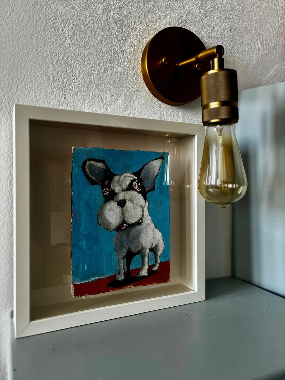 A framed painting of a dog with a large head and expressive eyes, set against a blue background, placed on a gray surface under a wall-mounted light fixture with a gold-colored base.
