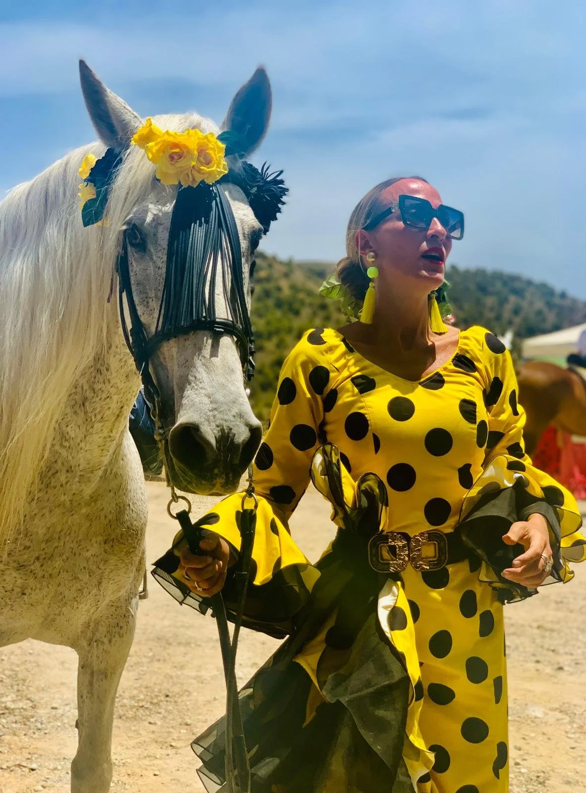 A woman in a traditional yellow Flamenco dress with black polka dots, large sunglasses, and yellow earrings stands beside a white horse decorated with yellow flowers on its head. They are outdoors under a blue sky with some clouds.