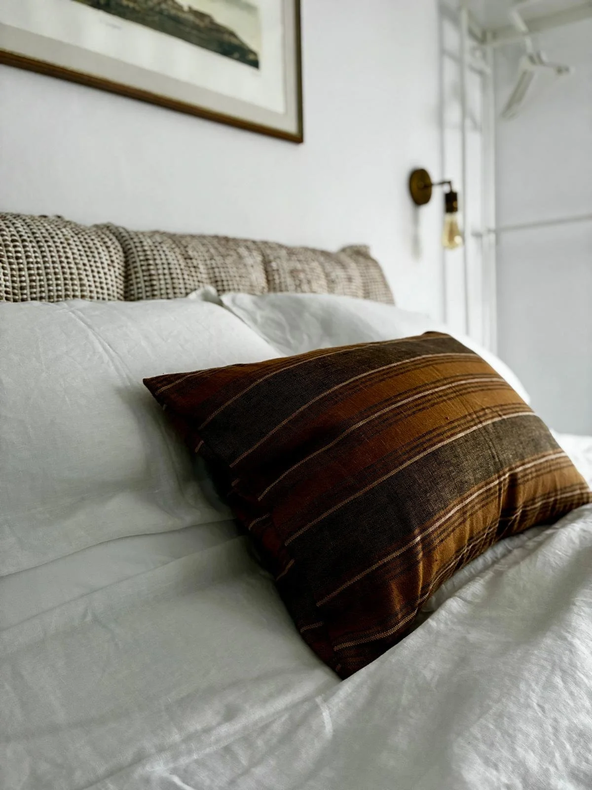 Close-up view of a bed with white sheets, a multi-colored brown striped pillow, and a headboard made of woven fabric pattern, in a minimalistic bedroom with white walls and framed artwork.