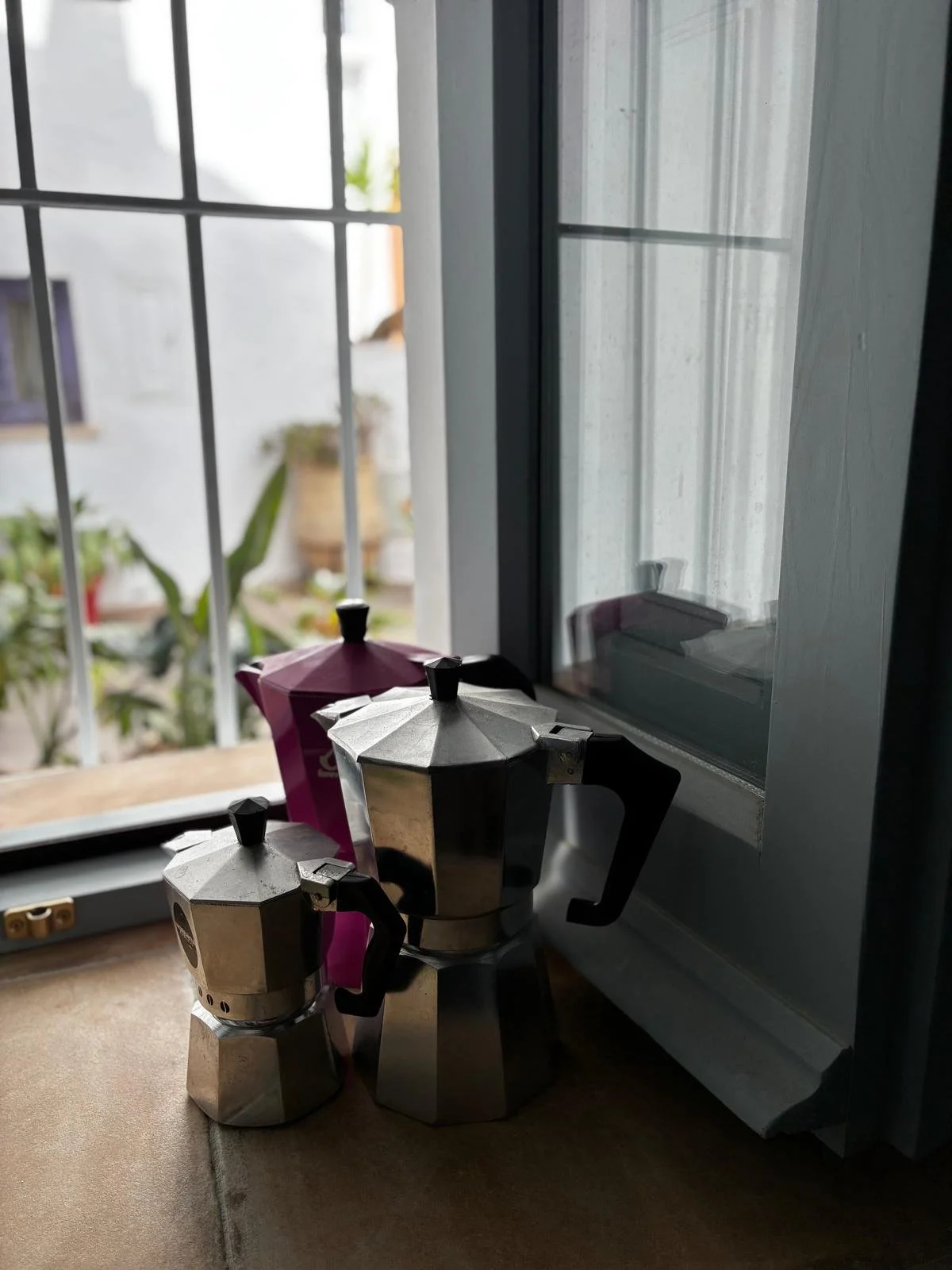 Close-up of three moka pots, two small and one large, on a kitchen windowsill with a window and potted plants outside.