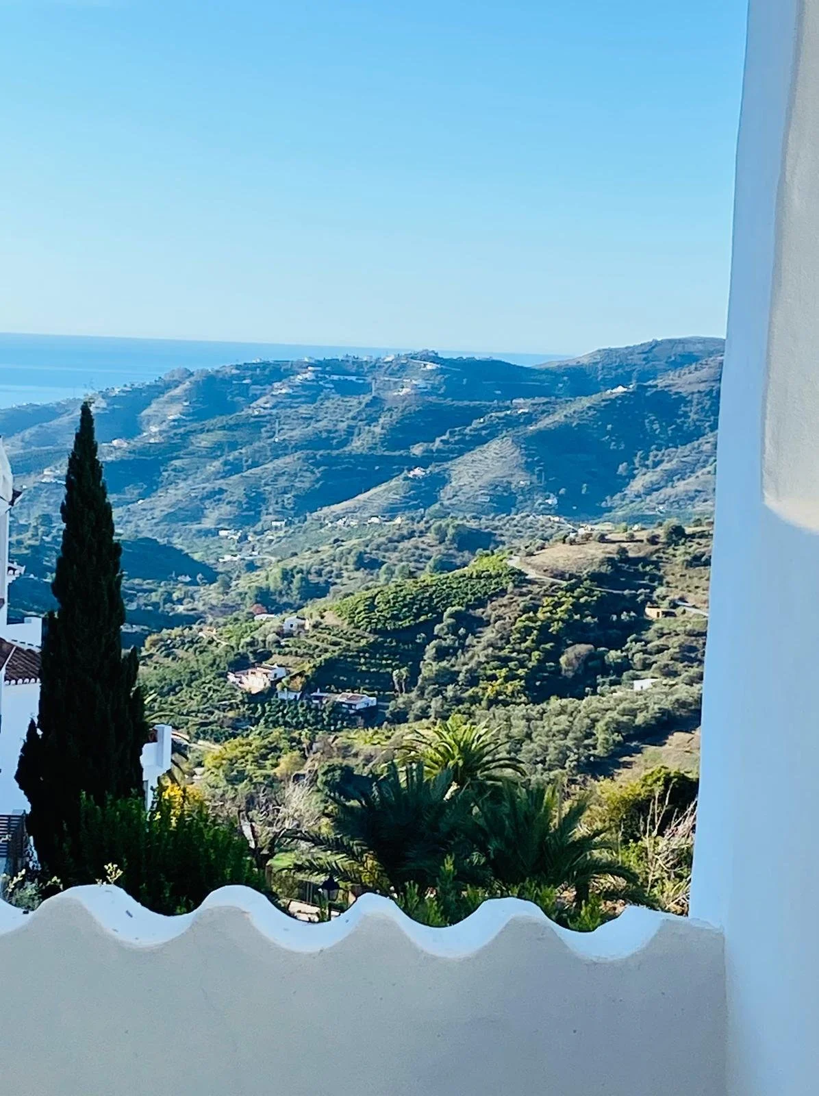 Scenic view from the chill-out area framed by white walls and a terrace. The view is of the hillside landscape with green vegetation, scattered houses, and mountains under a clear blue sky, with the sea in the background.