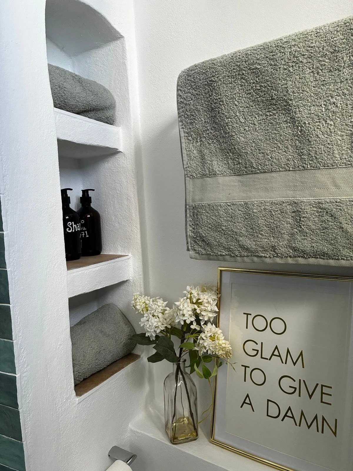Bathroom wall with stained glass shelves, gray towels, decorative framed quote, white flowers in glass vase, and brown bottles.