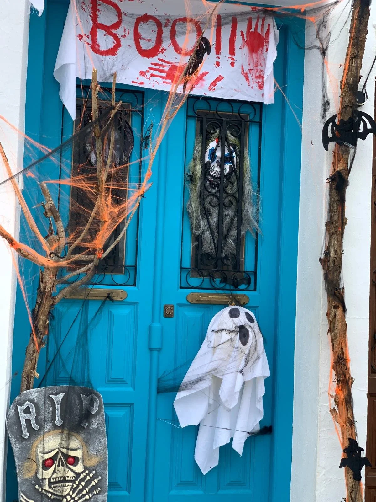 A blue door decorated for Halloween with a white sheet ghost, a tombstone, cobwebs, spider decorations, a banner with red handprints and the word 'BOO!!', spooky trees with cobwebs, and black bat cutouts.