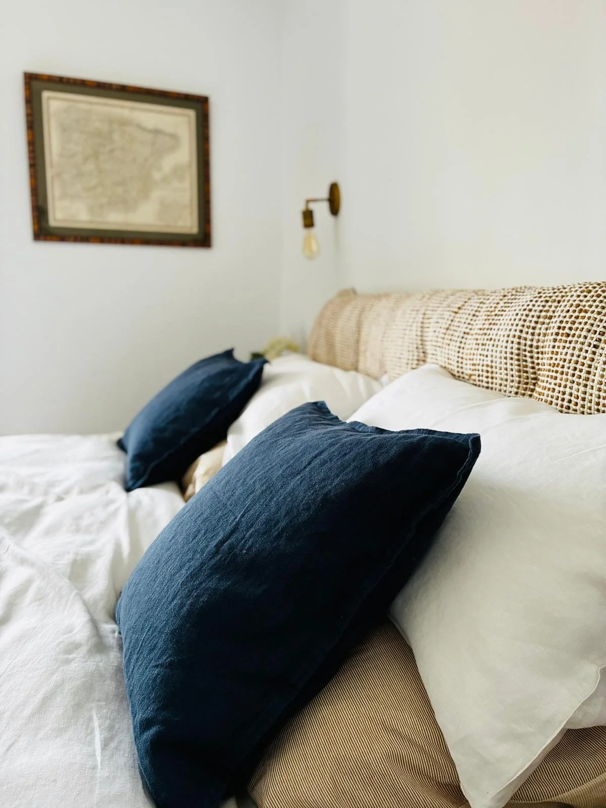 A neatly made bed with white sheets, navy blue pillows, a beige headboard, and a wall-mounted light fixture in a bedroom.