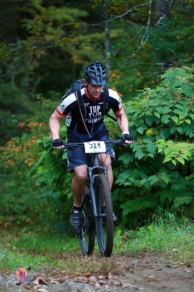 A man riding a mountain bike on a dirt trail through a wooded forest area, wearing a black helmet, black gloves, and a black and white cycling jersey.