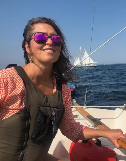 Woman with sunglasses on a boat, smiling, with sailboats in the background on the water.
