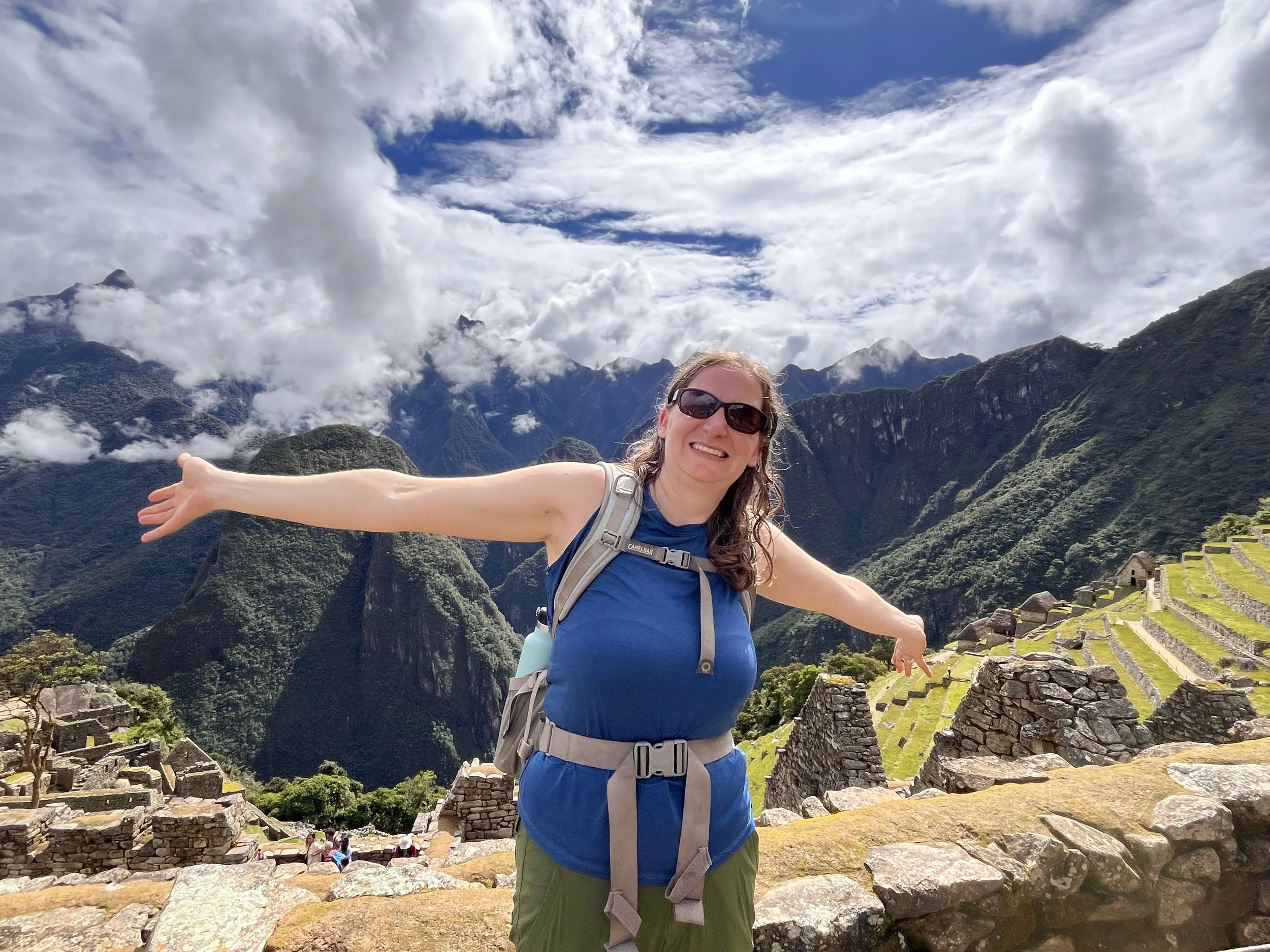 A smiling woman wearing sunglasses and a blue tank top with a backpack stands with arms outstretched amid ancient stone ruins at Machu Picchu, surrounded by lush green mountains and a partly cloudy sky.