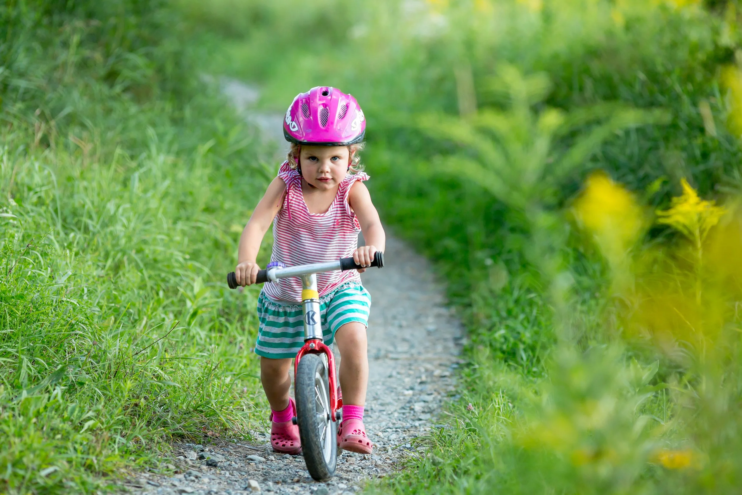 A young girl riding a bicycle on a narrow dirt and gravel path surrounded by green foliage and tall grass.