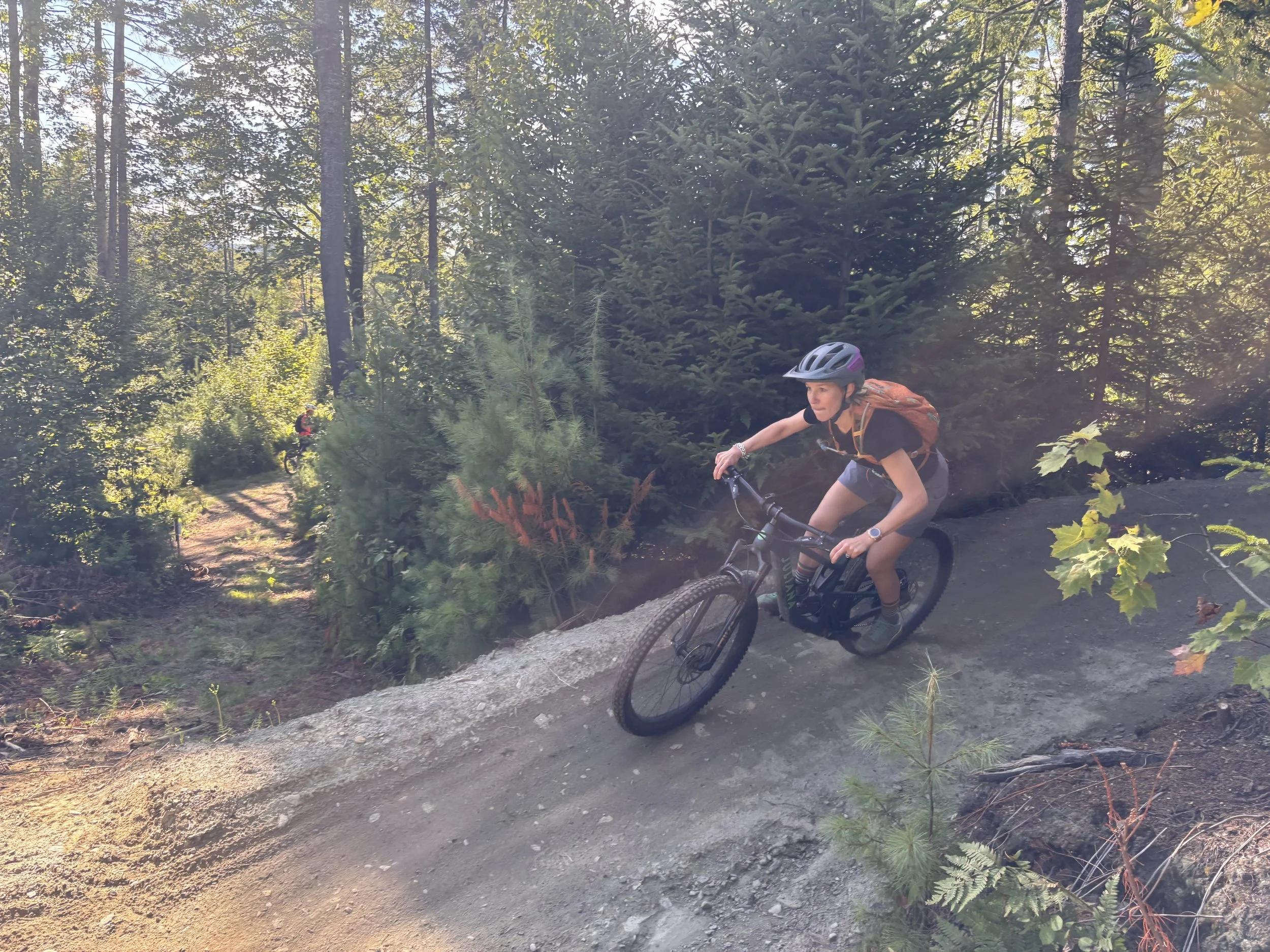 A woman riding a mountain bike on a dirt trail through a dense forest with tall trees and green foliage, wearing a helmet and backpack.
