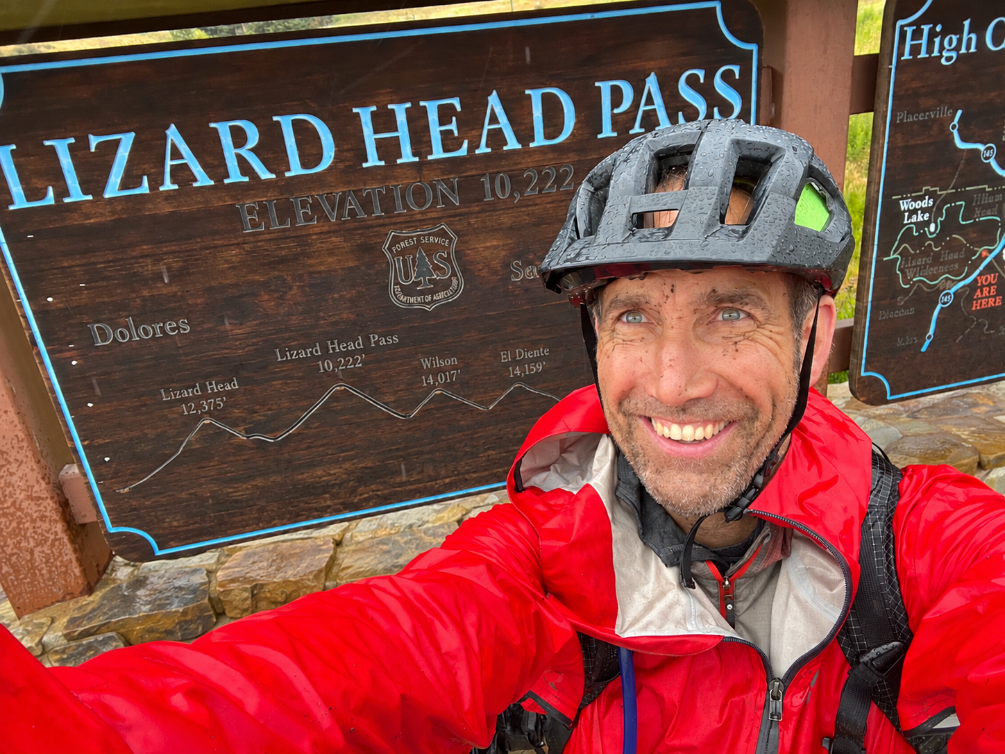 Smiling man in a red rain jacket and helmet taking a selfie at Lizard Head Pass, with a wooden sign showing elevation of 10,222 feet and a trail map in the background.