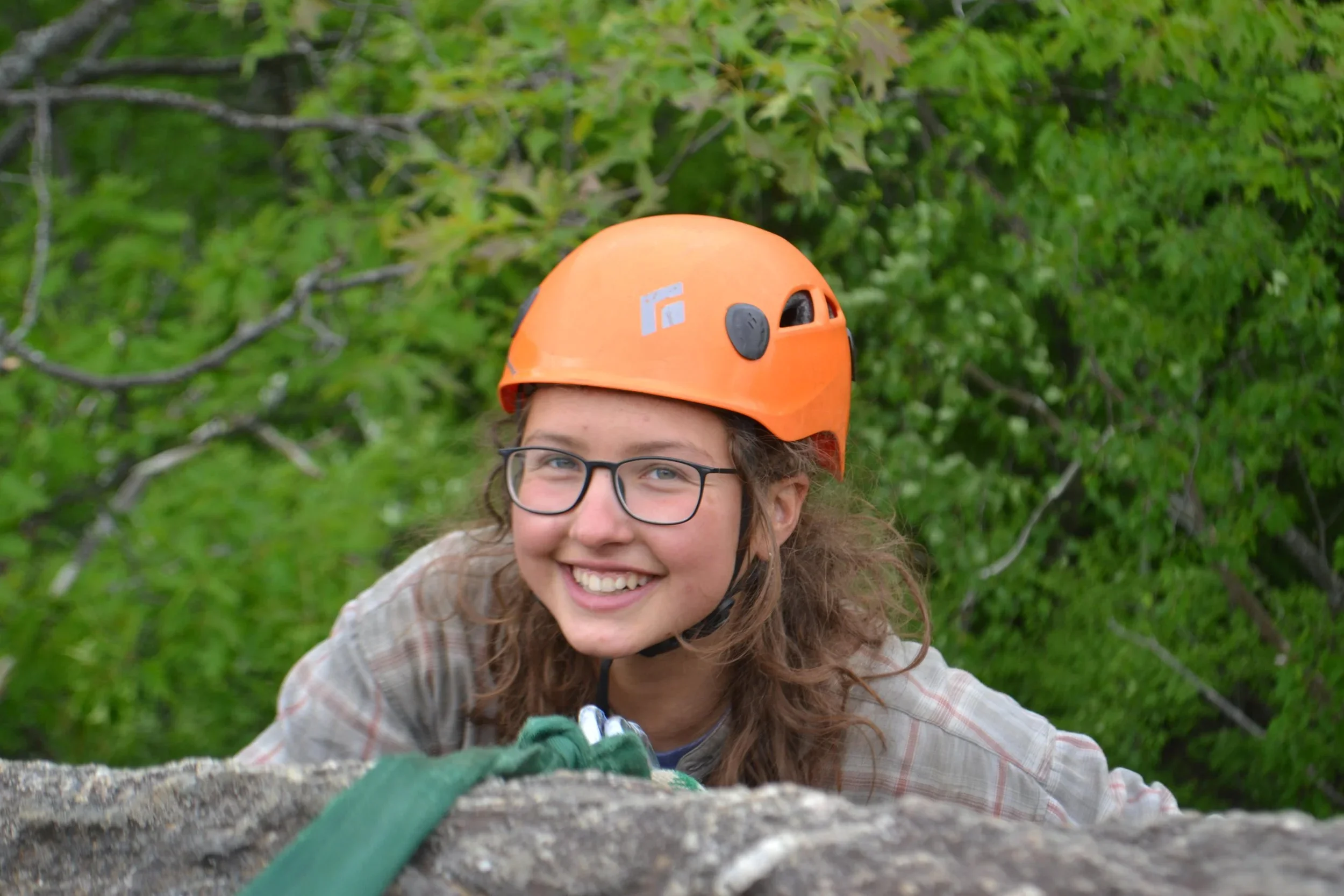 A young girl with glasses smiling while rock climbing outdoors, wearing an orange helmet and surrounded by green foliage.