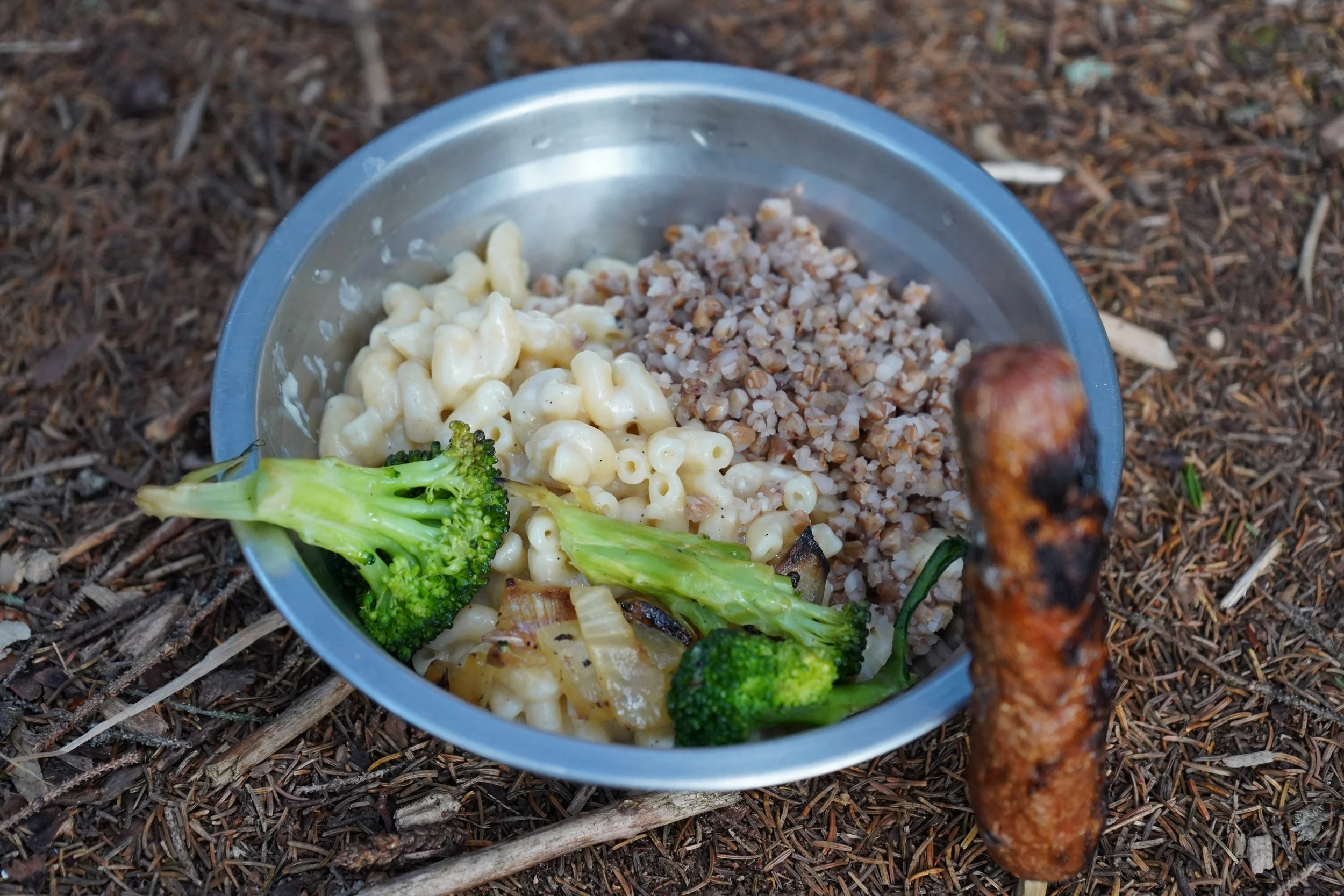 A metal bowl filled with macaroni and cheese, chopped cooked buckwheat, and broccoli florets, placed on a bed of pine needles and twigs outdoors, with a grilled sausage held vertically above the bowl.