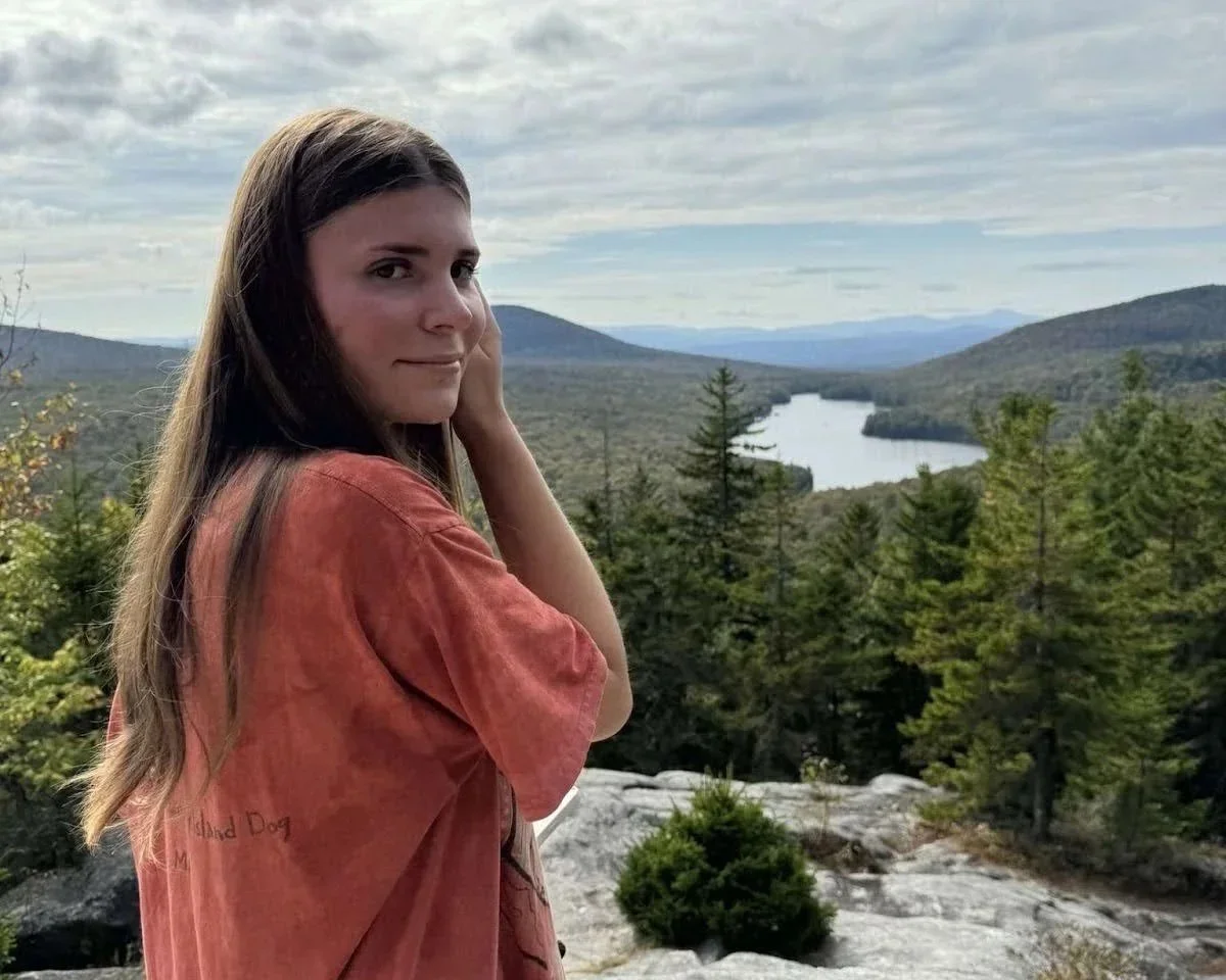 Young woman with long brown hair standing outdoors near trees, overlooking a scenic view of a river or lake in a valley with mountains in the distance under a cloudy sky.