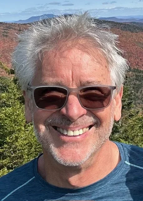Smiling man wearing sunglasses outdoors with a scenic mountain landscape and trees in the background.
