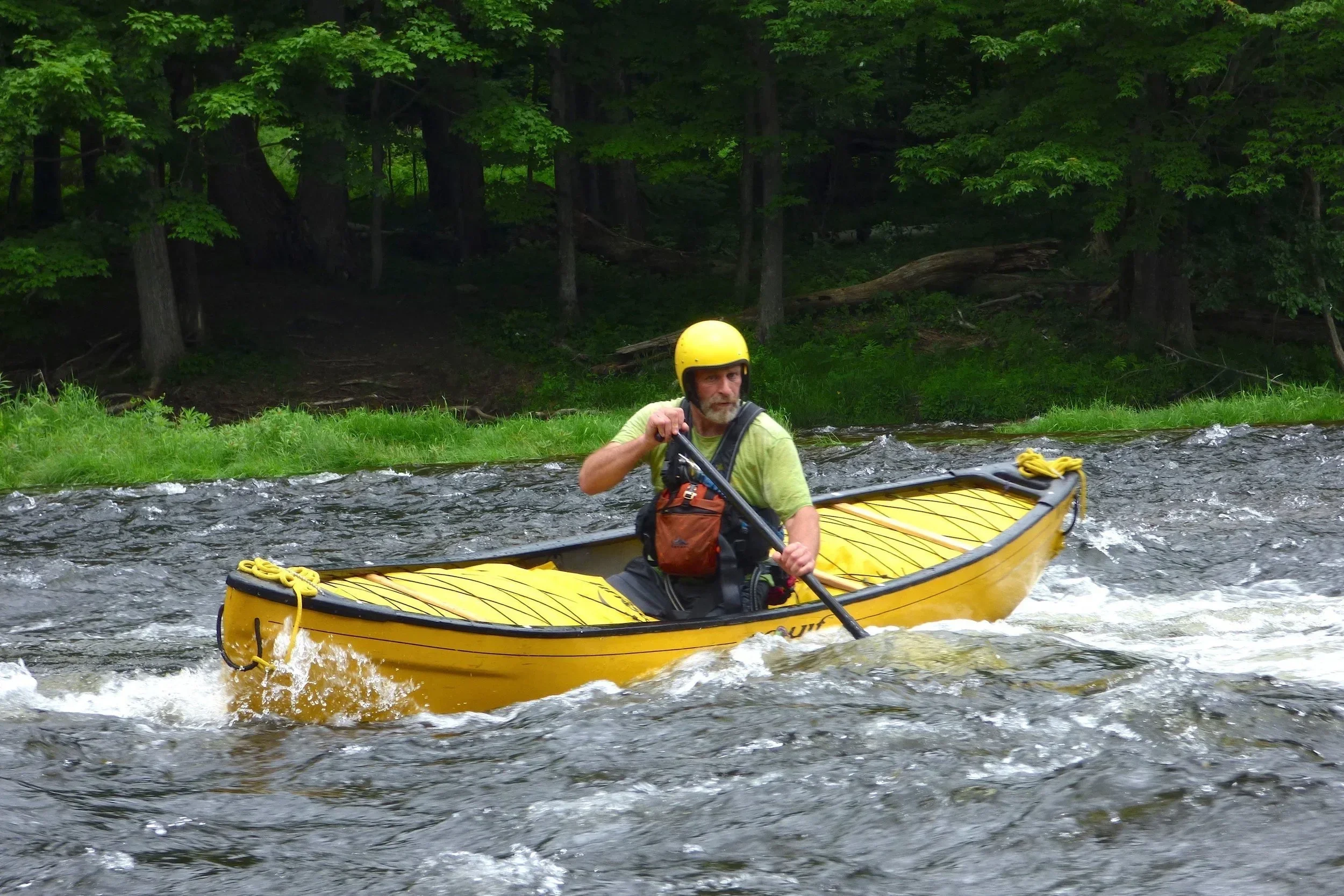 A man kayaking on a river with a lush green forest in the background, wearing a yellow helmet and a green shirt.
