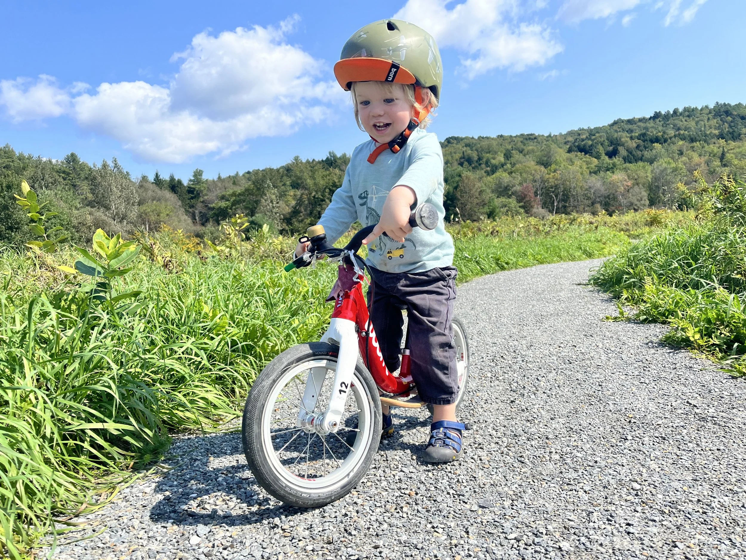 A young child with blonde hair smiling while riding a red and white bicycle on a gravel path in a rural area with green hills and blue sky.