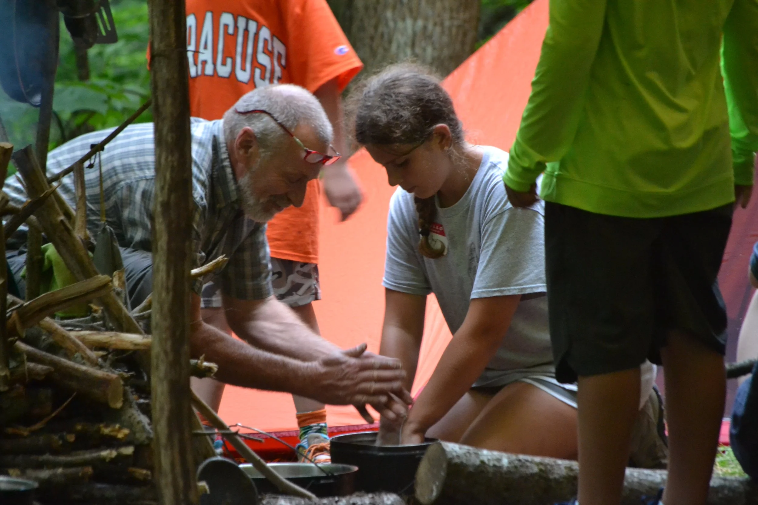 An older man and a young girl are engaged in a hands-on activity likely outdoors, surrounded by trees and wooden branches, with a group of people nearby, some wearing bright clothing.