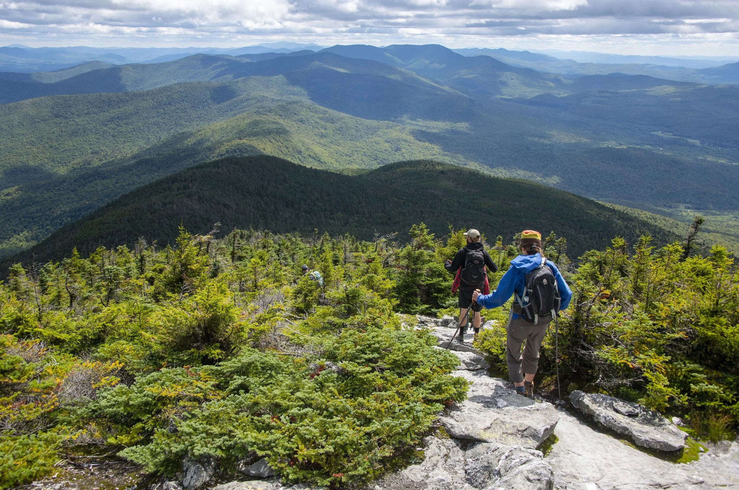Three hikers walking along a rocky trail through green mountain forest with layered mountain ranges in the background under a partly cloudy sky.
