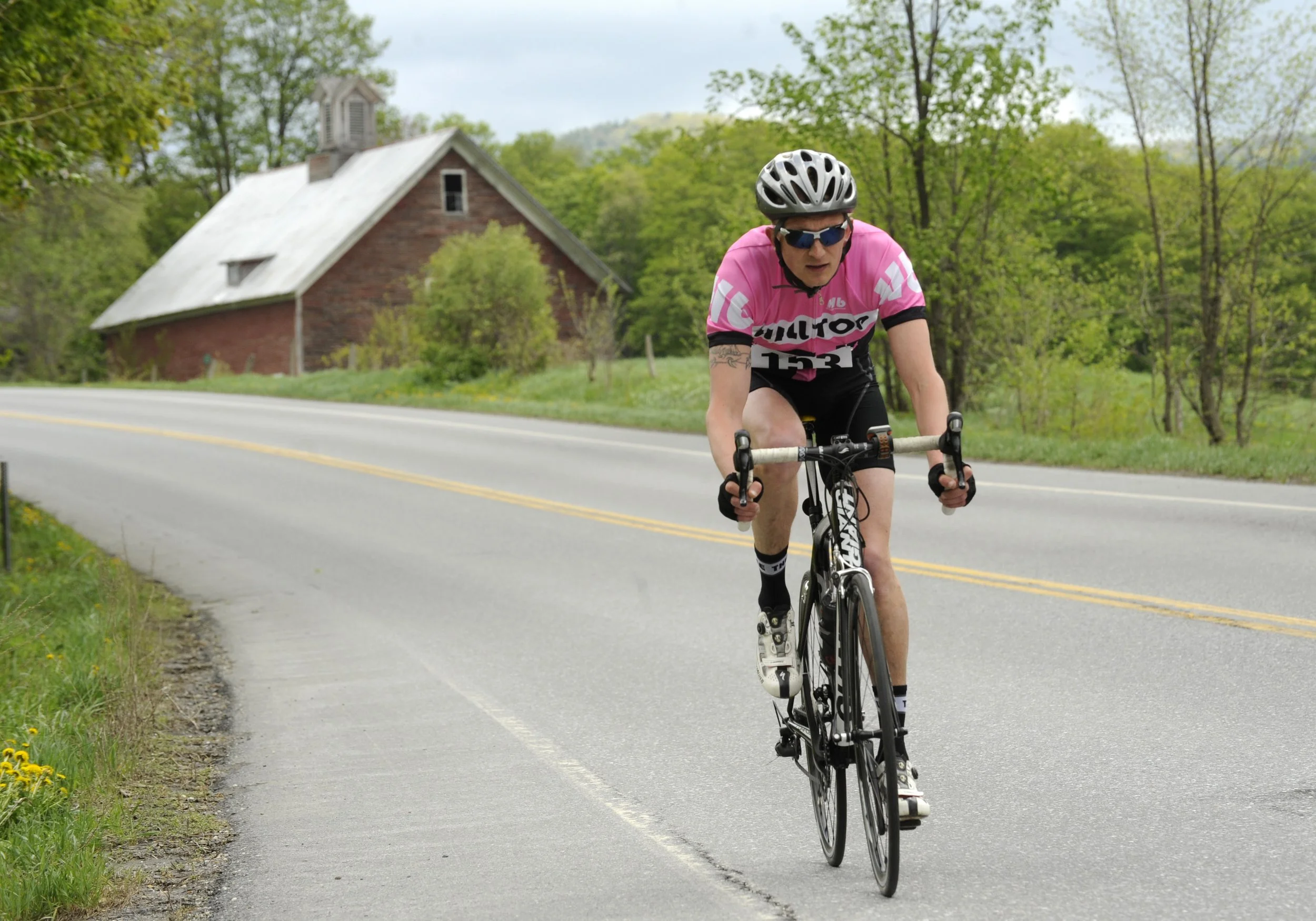 A man wearing a pink cycling jersey, black shorts, sunglasses, and a helmet, riding a bicycle on a rural road with a church and green trees in the background.