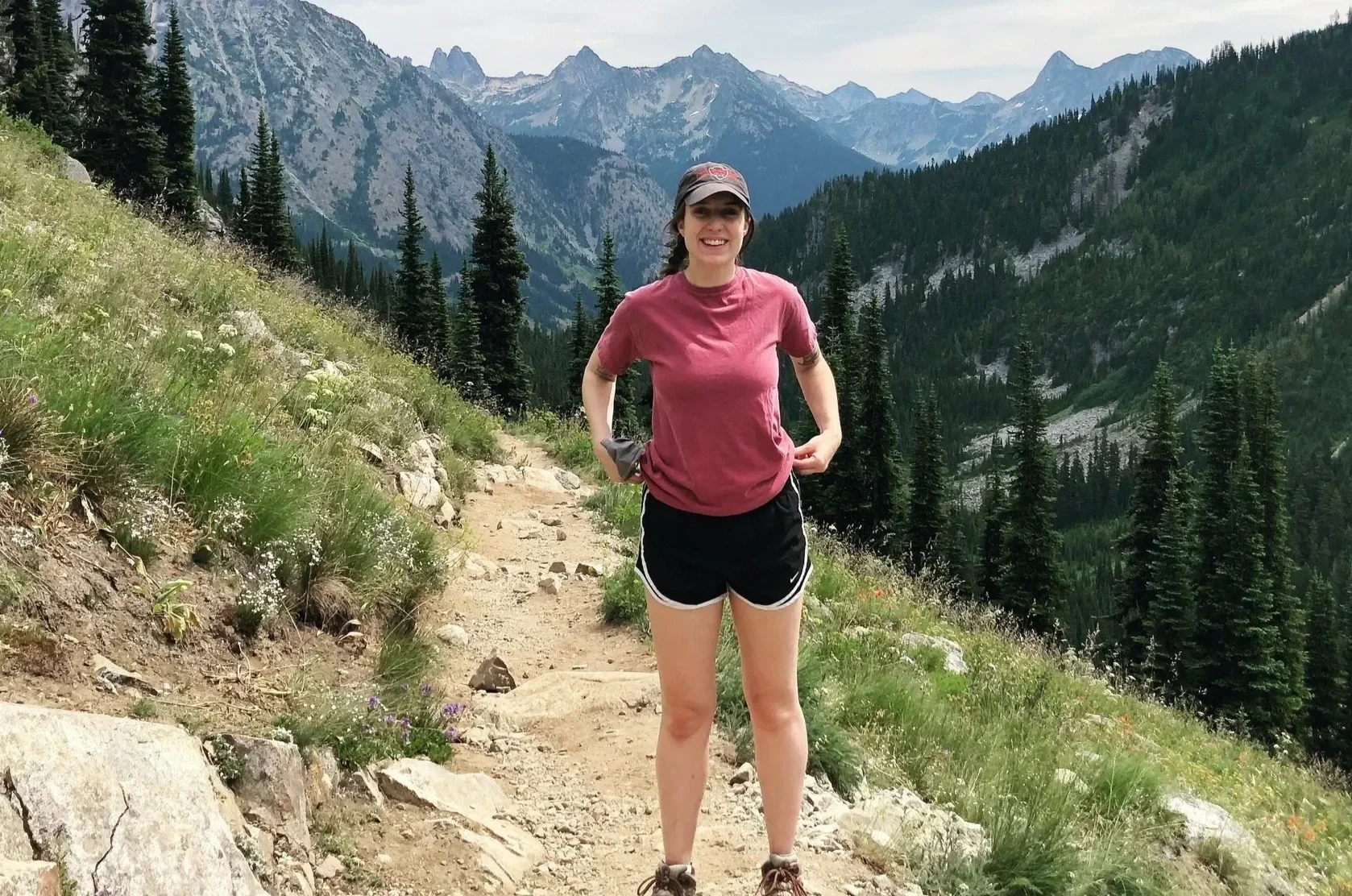 A woman in a pink t-shirt and black shorts standing on a mountain trail surrounded by tall pine trees, with a mountain range in the background.
